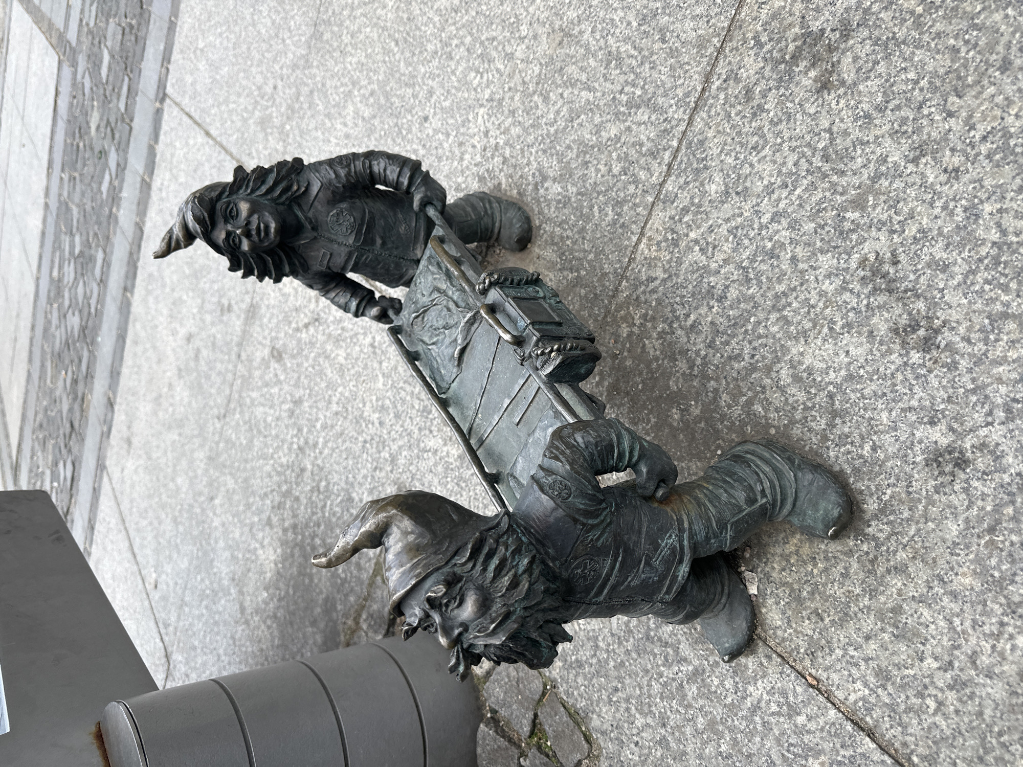 The image depicts two bronze dwarf statues in Wrocław, Poland. They are positioned on a gray stone surface with a concrete curb visible on the left. The dwarves are hauling a sled carrying a chest. Both dwarves are wearing pointed hats and detailed clothing. The scene suggests a playful, whimsical element typical of Wrocław's dwarf statues.