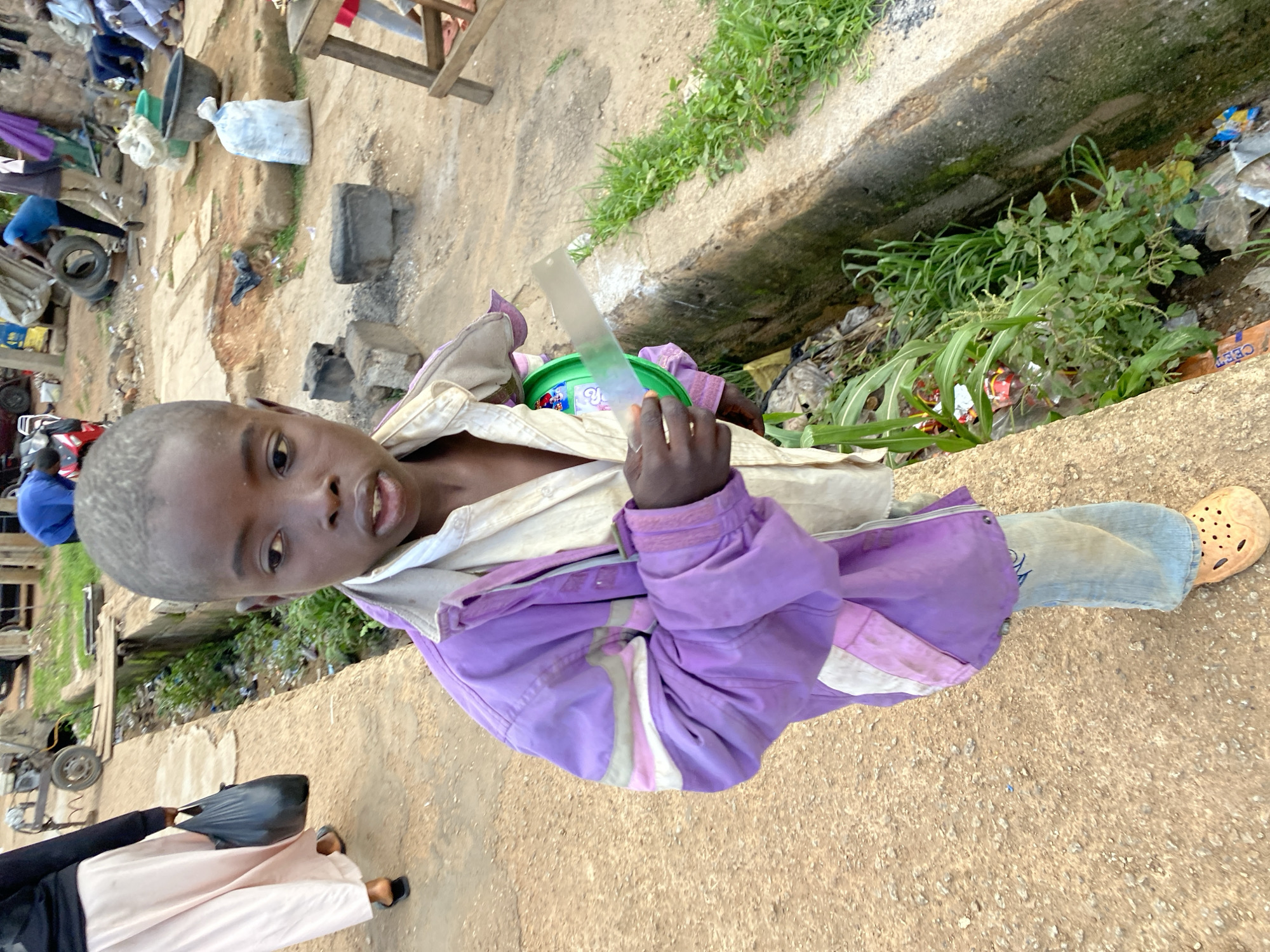 The image depicts a young boy in Bukuru, Nigeria, standing next to a drainage ditch filled with debris and vegetation. He is wearing a light-colored shirt and a purple jacket with white and pink accents, along with light blue jeans and orange Crocs. In his right hand, he holds a green object and a clear, flat piece of plastic or glass, possibly a ruler. The background shows a dirt pathway, a partially visible woman carrying a black bag, and various objects and people in the distance, suggesting a busy, informal market or street scene.