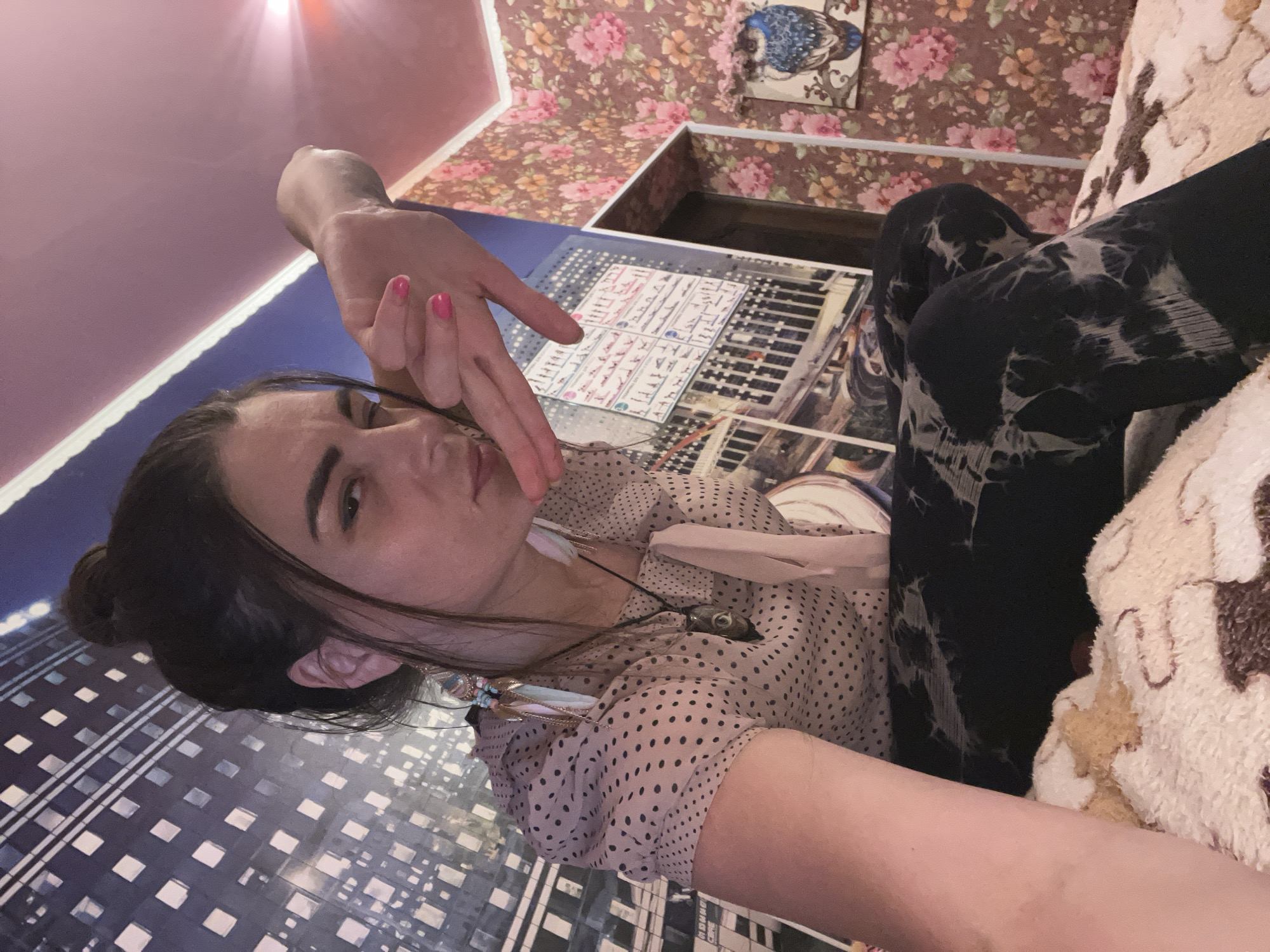 A young woman with dark hair pulled into a bun is seated on a bed, her body angled towards the camera. She is wearing a light-colored, polka-dotted blouse and dark, patterned leggings. Her right hand is raised, with her fingers extended and touching her chin. She has bright pink fingernail polish. Her left arm is resting on her lap. The background consists of wallpaper with a floral pattern on the right and a large mural depicting a cityscape on the left. A framed picture of a blue owl is visible on the wall to the right. The overall lighting is warm and appears to be artificial. The location is identified as Krasnoe-na-Volge, Russia.