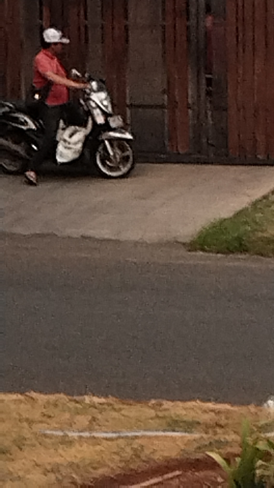 A person wearing a red shirt and a white cap is sitting on a scooter. The scooter is black and white, with a white bag attached to the front. The person is facing to the left, and their hands are on the handlebars. The scooter is parked on a paved surface, in front of a wooden fence. In the background, there are some green plants. The overall scene appears to be in a residential area.