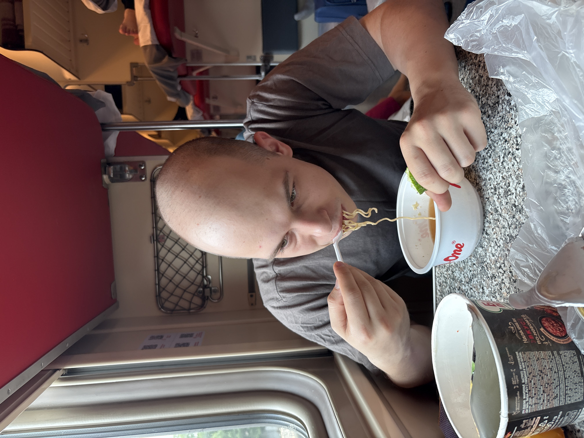 The image depicts a young, bald man eating instant noodles from a white bowl with a plastic fork while seated at a table, likely inside a train compartment. The compartment appears to be in Russia, possibly Gryady, given the context. The man is wearing a grey t-shirt. In the background, there are visible train bunks, curtains, and other passengers, including a person whose feet are visible on a bunk. There are two containers of instant noodles on the table, one partially open. The overall scene suggests a meal being consumed during travel.