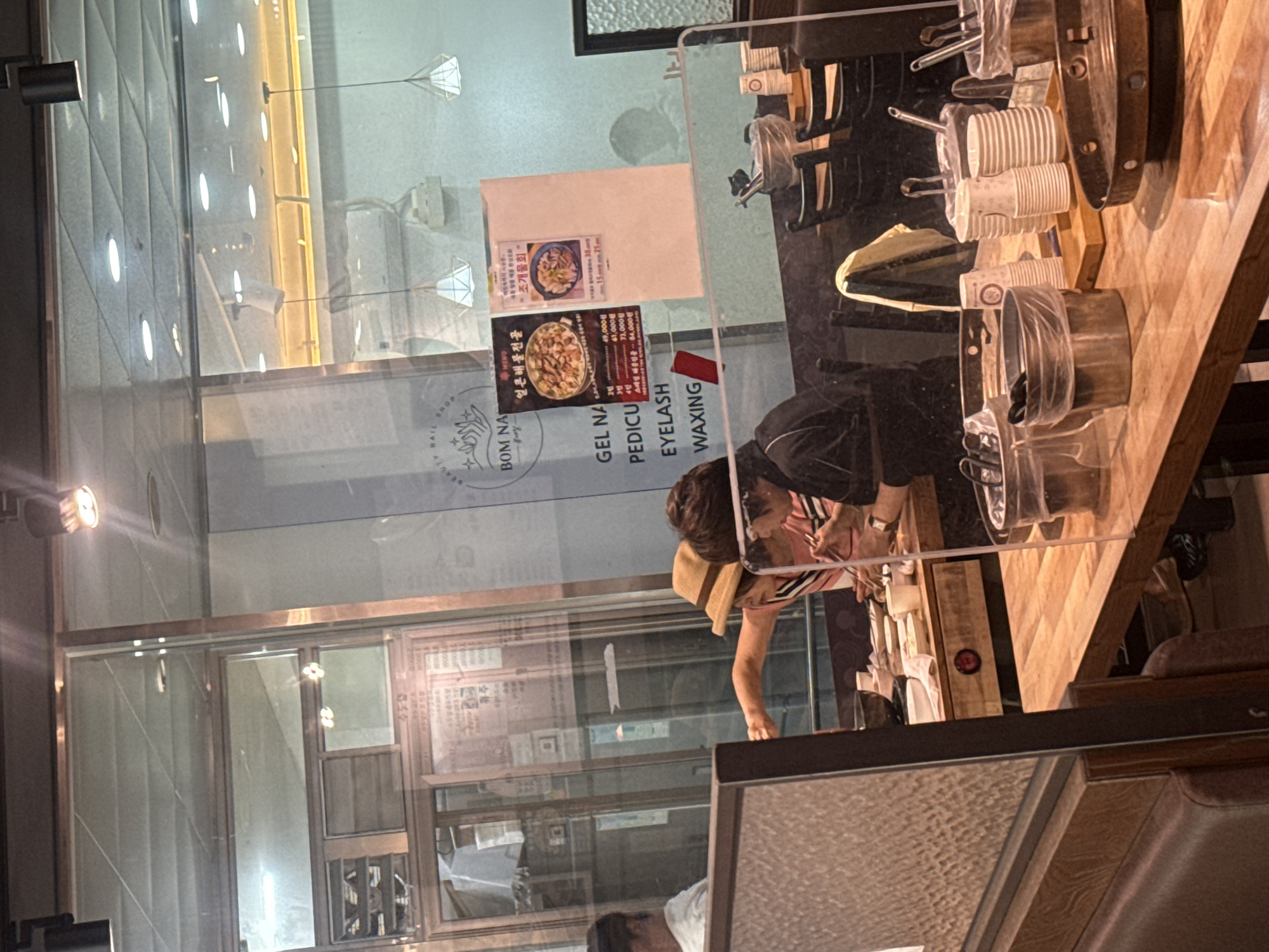 Here is a description of the image:
The image shows the inside of a business establishment, likely a nail salon or beauty shop named "BOM NA," located in Gimpo-si, South Korea. The view is partially obstructed by a transparent partition. Posters displaying Korean text and food images are visible on the windows. In the foreground, two people are visible. One wears a straw hat and a striped shirt, while the other wears a dark jacket and appears to be interacting with the customer. Cups and salon equipment are on the right-hand side. Outside the store, there is a building with a patterned facade and lit with numerous lights.