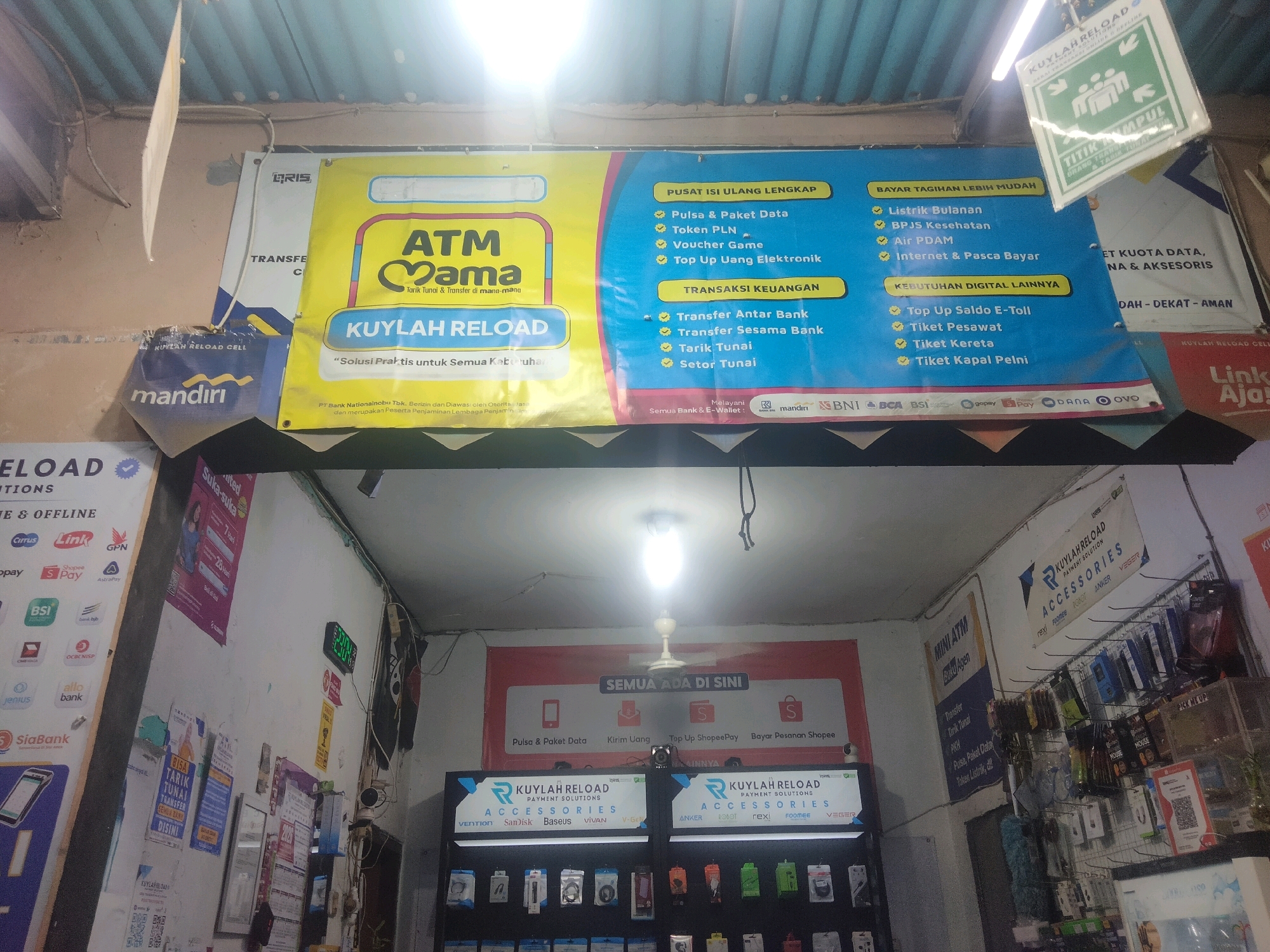 The image shows the exterior of a small shop, likely in Suka Sari, Indonesia, with a large banner advertising "KUYLAR RELOAD PAYMENT SOLUTIONS". The banner lists various services including ATM transfers, bill payments, and digital needs such as top-ups and ticket purchases. Several logos of Indonesian banks and e-wallets are visible on the banner, indicating partnerships. Below the banner, the shop appears to sell mobile accessories, with displays showcasing various brands like Vention, Baseus, Remax, Anker, and Veger. The shop is open for business, with products displayed both on shelves and in hanging racks. A ceiling fan is visible, and the lighting suggests it is daytime or the shop is well-lit. The overall scene depicts a typical Indonesian convenience store or payment point offering a range of services and products.
