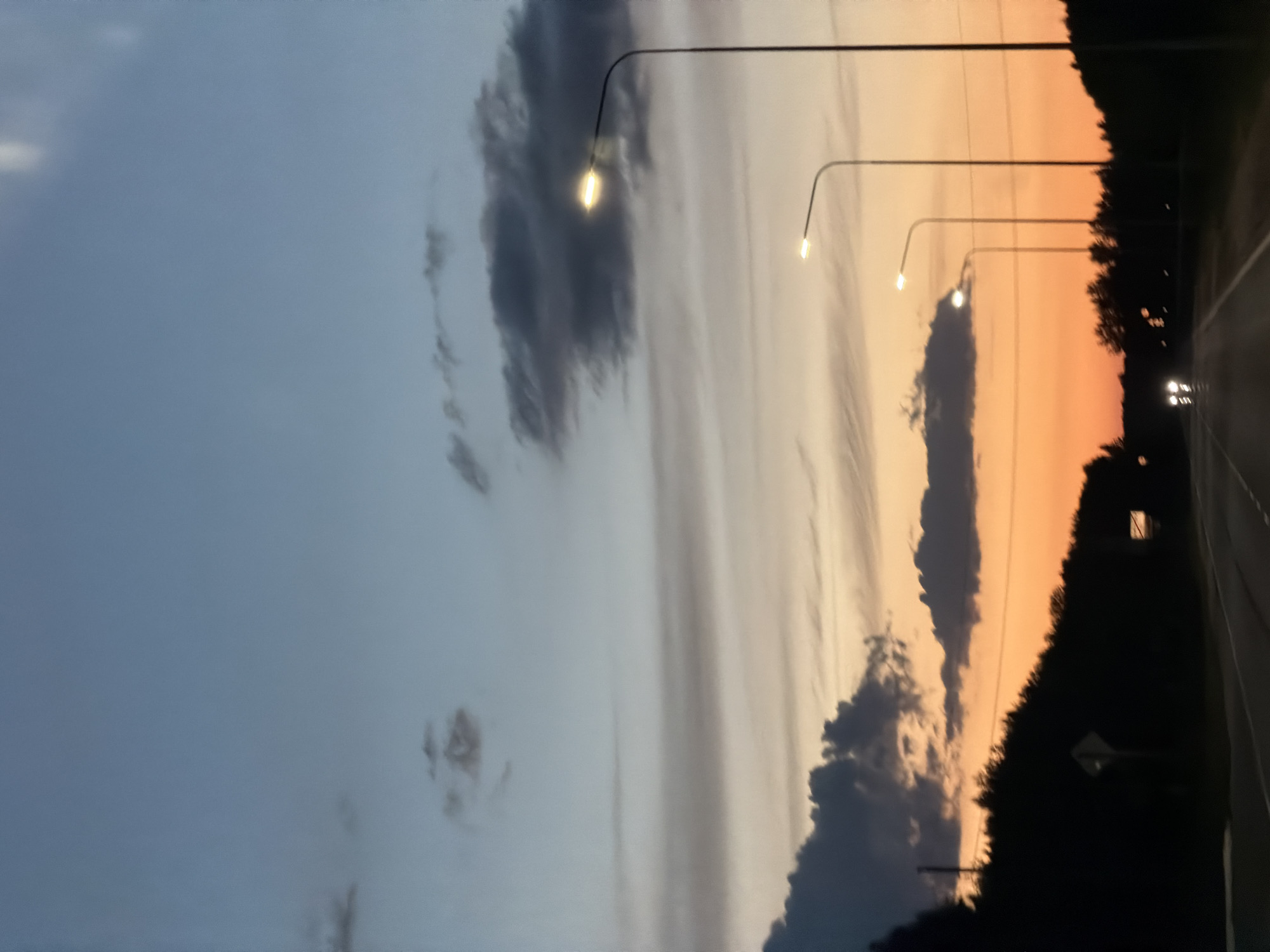 The image depicts a road at dusk, likely in or near the city of Laishevo, Russia. The sky exhibits a gradient of colors, transitioning from a light blue at the top to hues of orange and yellow near the horizon, indicating sunset. Several clouds are visible, some dark and dramatic, others wispy and stretched. Streetlights are illuminated along the side of the road, casting a soft glow. Silhouetted trees and vegetation line the edges of the road, creating a dark boundary against the lighter sky. In the distance, headlights from an approaching vehicle are visible on the road. The scene appears to be a quiet, transitional moment between day and night.