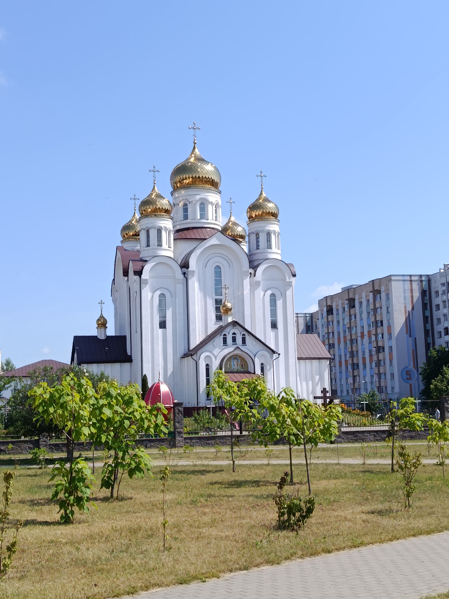 A bright, sunny day is captured in this image, with a clear blue sky dominating the upper portion. The main subject is a large, white Orthodox church with several golden domes, each topped with a cross. The church has intricate architectural details, including arched windows and decorative moldings. In front of the church, a smaller attached structure features a mosaic above its entrance.
The foreground is occupied by a neatly maintained lawn, with several young trees and shrubs planted in rows. A low, dark fence encloses the area around the church. In the background, to the right of the church, stands a tall, modern apartment building with a colorful facade featuring vertical stripes of blue, orange, and grey. A large blue sign with a white number "5" is visible on the building.
The overall impression is one of a peaceful, well-kept urban setting. The bright sunlight suggests it is daytime, likely late morning or early afternoon. No people are visible in the frame, and no specific event appears to be taking place. The scene conveys a sense of tranquility and architectural beauty.