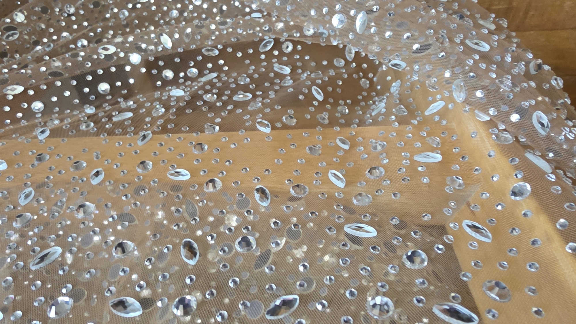 A close-up image showcases a piece of translucent, light brown or beige fabric densely embellished with sparkling clear rhinestones. The sheer material, possibly a mesh or tulle, is draped, creating gentle undulations and soft folds across the frame.
Numerous rhinestones of varying shapes and sizes are scattered across the fabric. Many are small and circular, while others are larger, often oval or marquise-shaped with faceted surfaces that catch and reflect light, creating a brilliant, glittering effect. The rhinestones are irregularly distributed, adding texture and shimmer to the textile.
Through the sheer fabric, a blurred, warm medium-brown background is visible, suggesting a wooden surface such as a table or floor. No people or specific activities are depicted; the image is a static portrayal of the embellished fabric. There is no visible text. The lighting is bright, emphasizing the reflective qualities of the rhinestones. The provided location context is Blagoevgrad, Bulgaria, but no visual cues in the image itself indicate this location.