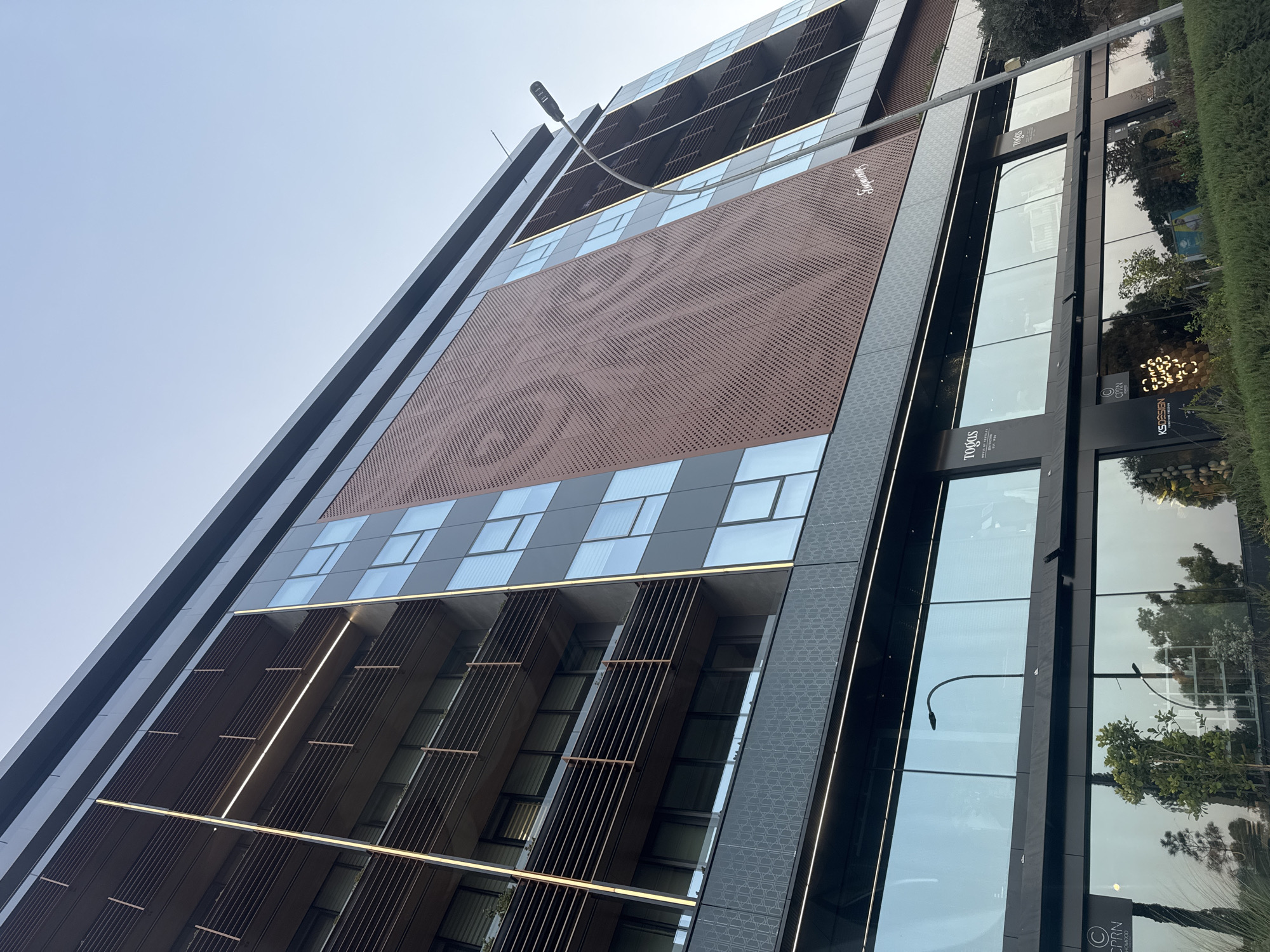 A low-angle, wide shot captures a modern, multi-story building in Agios Athanasios Municipality, Cyprus, under a clear, light blue sky. The building features a dark facade with prominent horizontal balconies, each protected by dark metal railings and vertical slats. Integrated vertical lighting strips accent the building's edges. A large section of the facade is covered in a reddish-brown perforated metal paneling arranged in a patterned grid. The ground floor of the building consists of large, reflective glass windows displaying the reflections of trees and the sky, with visible signage including "TOOGAS HOUSE OF DESIGN EST 1998," "CPRRN HOME D," and "KSDESIGN." A tall, grey street lamp with a curved arm is positioned to the right of the building, extending upwards into the sky. The foreground shows a neatly trimmed green hedge.