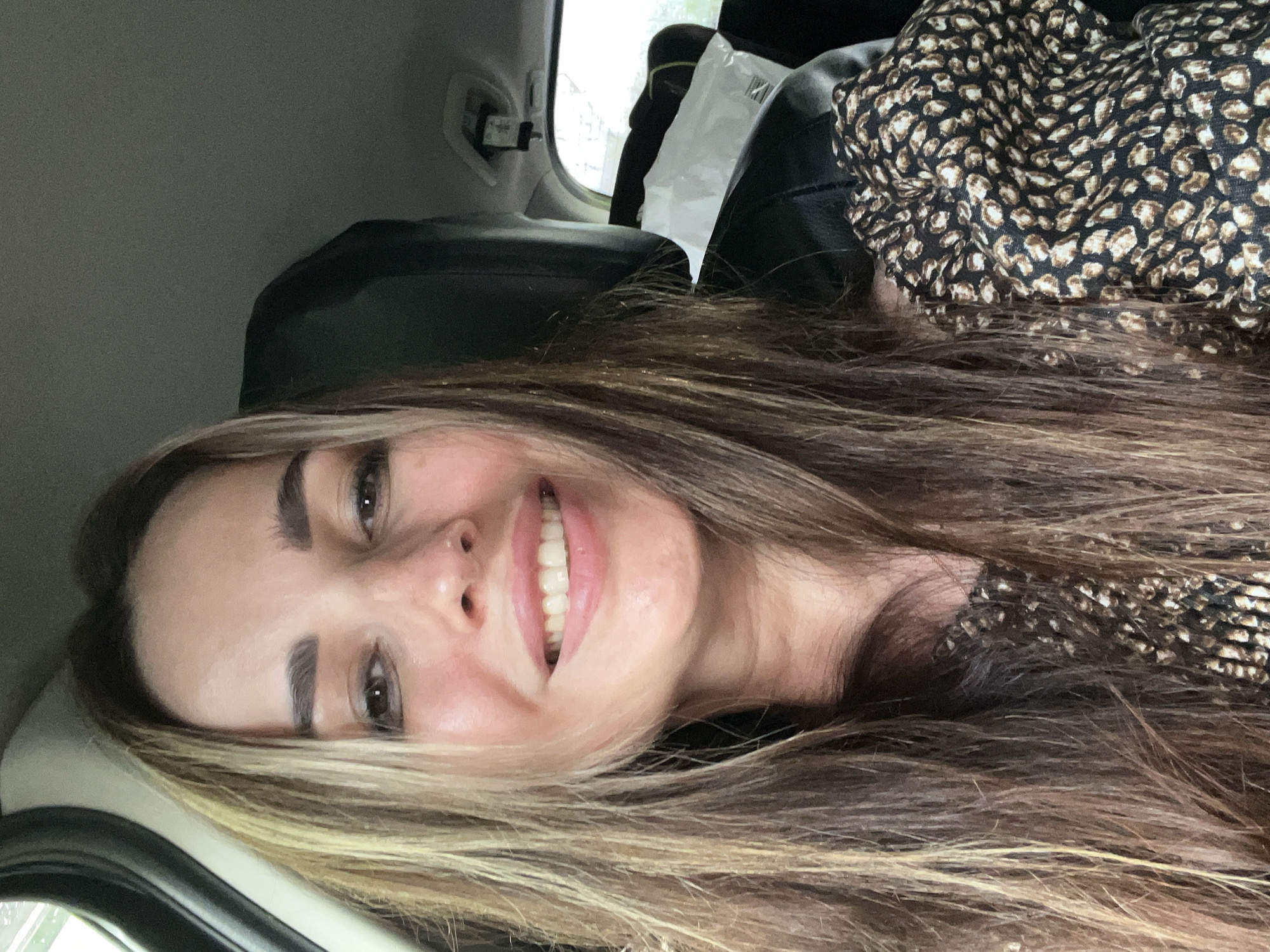 A woman with long, wavy brown and blonde-highlighted hair smiles at the camera in a selfie taken from inside a vehicle. She is wearing a patterned top. The background shows the interior of the car, including a headrest and a window with a blurred view of the outdoors.