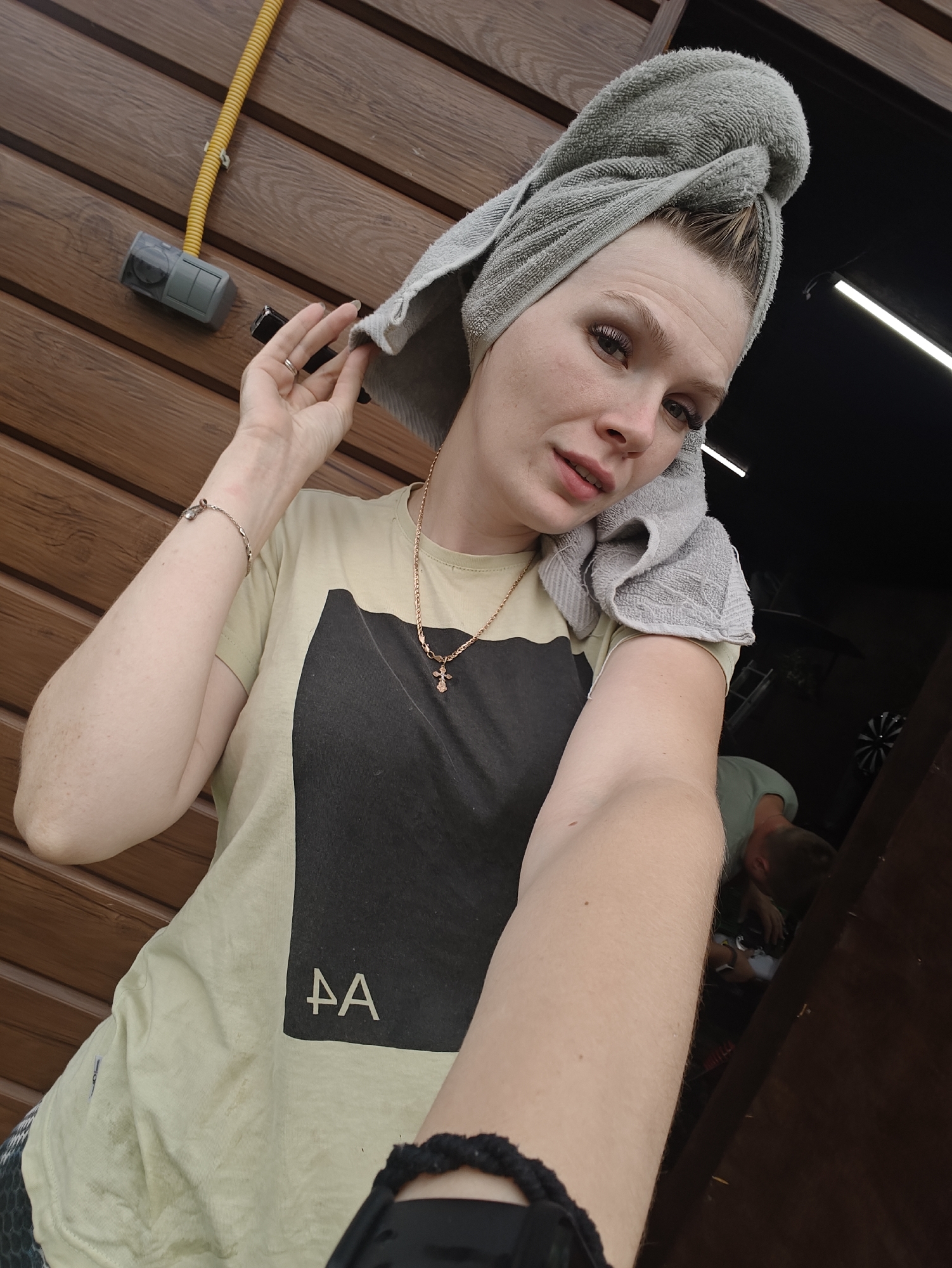 A young woman with a towel wrapped around her hair and a towel in her right hand is taking a selfie. She is wearing a light yellow t-shirt with a black graphic design. She has on a necklace with a cross pendant and a bracelet on her left wrist. The background is a wooden wall with a yellow pipe and a grey electrical box attached to it. To the right, a dark opening reveals another person wearing a green shirt, hunched over. The scene appears to be outdoors, possibly in an urban or suburban setting. The location is reported as Russky Vozhy, Russia.