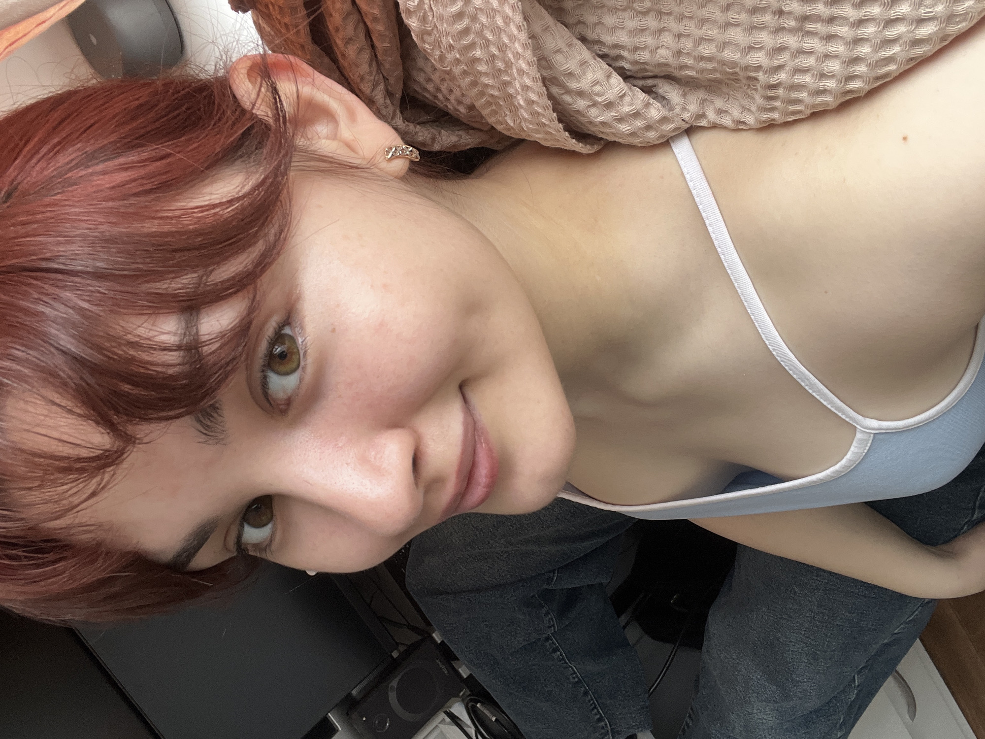 A young woman with auburn hair and hazel eyes is pictured indoors. She wears a light blue tank top with white straps and dark wash denim jeans. A textured beige shawl or blanket is draped over her shoulders. An earring is visible in her left ear. To the left of the frame is a black object, potentially a monitor or part of a computer setup, along with a small black speaker. The location and city information cannot be confirmed as there is no indication of location in the image provided.