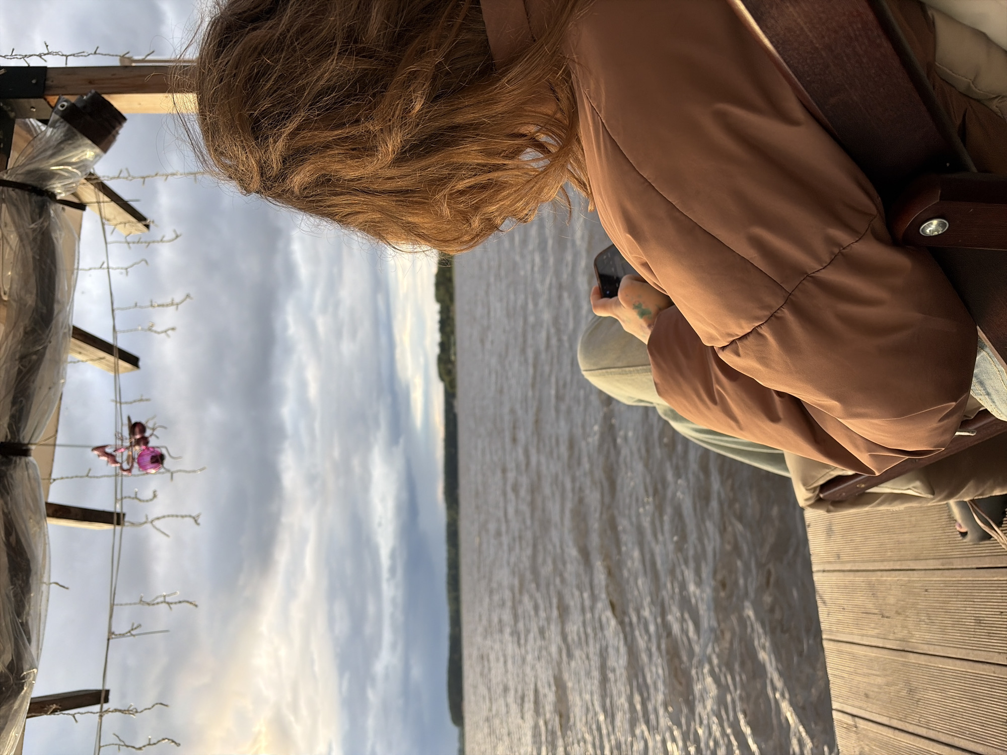 A person with auburn hair, wearing a brown puffer jacket and light-colored pants, is seated on a wooden deck, looking out at a body of water. The person is holding a smartphone in their right hand. The water appears to be a lake or river, with a treed shoreline in the distance under a cloudy sky. Above the person, there is a canopy structure with lights and a pink decorative ornament hanging from a string. The scene is likely outdoors, possibly in a recreational area. The presence of the lake and treed shoreline suggests a natural setting. The wooden deck indicates a built structure for access to the water. The lighting suggests it is either early morning or late afternoon.