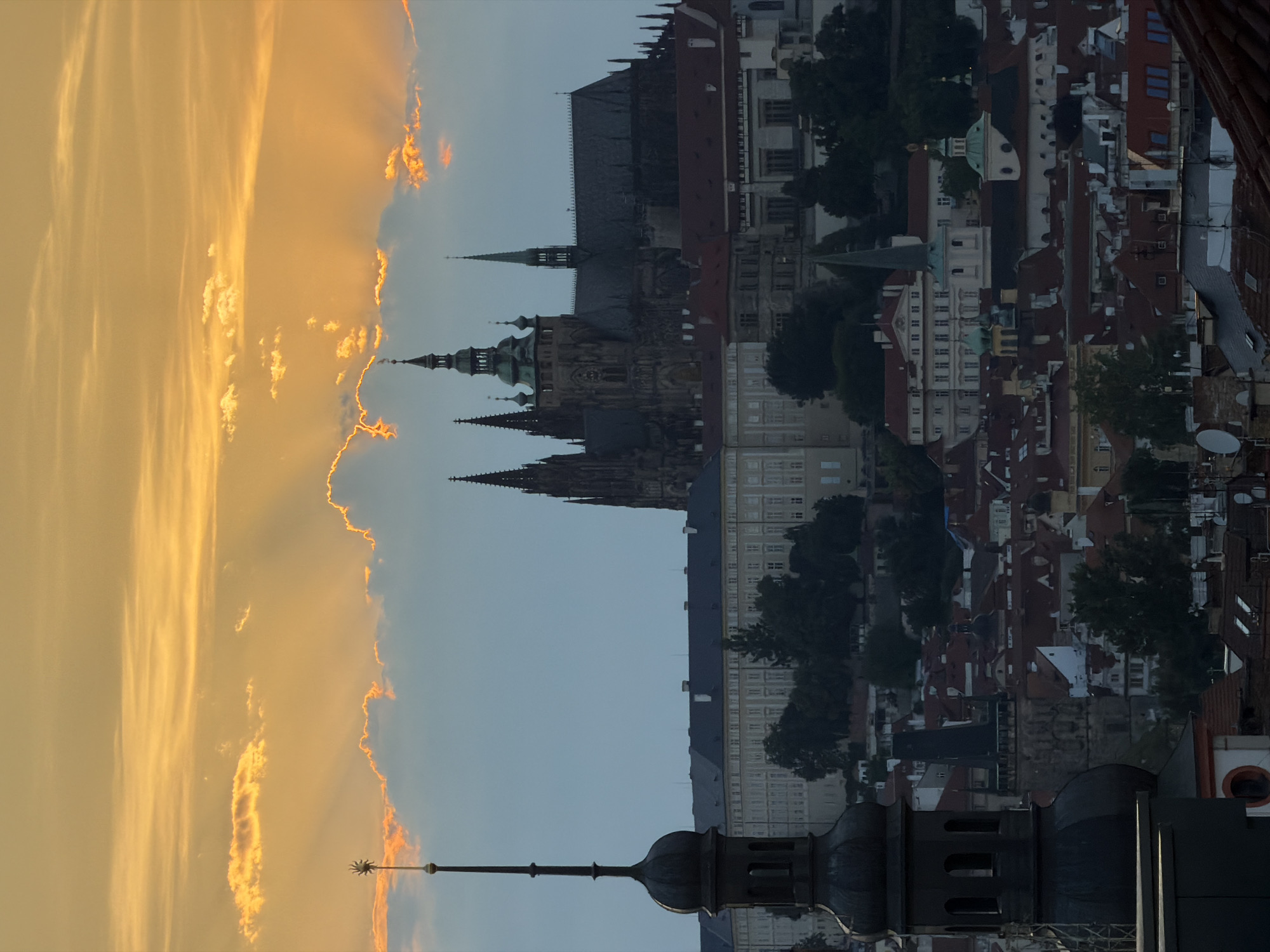 This is an aerial, vertical, medium shot of Prague, Czechia at dusk. The sky is filled with dramatic, illuminated clouds, with rays of sunlight breaking through. In the midground, a vast architectural complex, believed to be Prague Castle, dominates the skyline. Its Gothic spires and intricate stonework are silhouetted against the twilight sky. In the foreground, the dark, imposing structure of a church tower with a pointed spire, topped with a star ornament, commands attention on the left side of the frame. The lower portion of the image reveals a dense cityscape of red-tiled roofs and smaller buildings, indicative of a historic European city. No people are visible in the image.