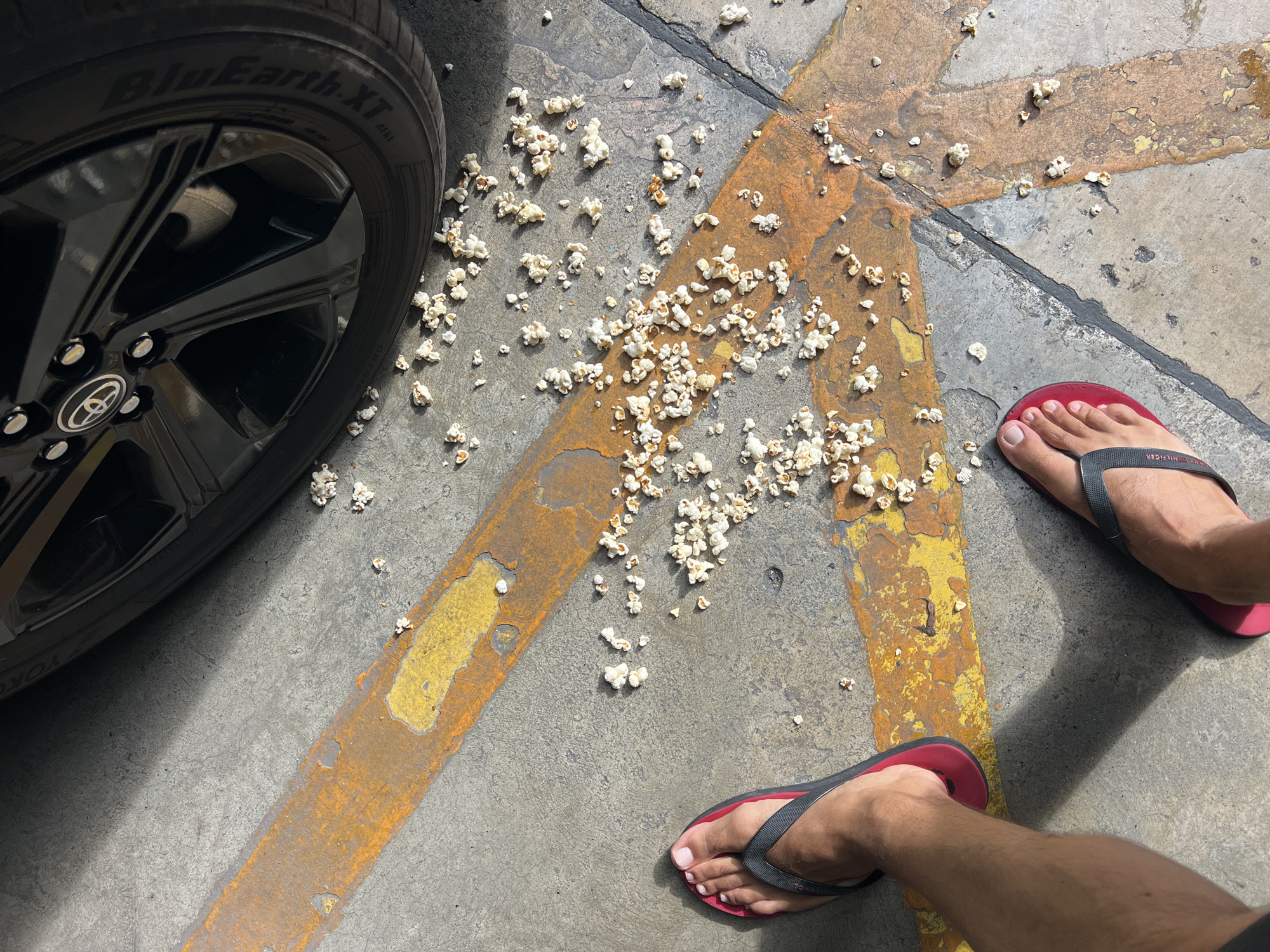In a parking lot area at Ban Rang Rak, Thailand, a scattering of popcorn is visible on a concrete surface marked with weathered yellow and orange lines. A black car wheel with a silver rim is positioned in the upper right portion of the frame. Two individuals' feet, wearing flip-flops, are in the lower half of the image, one with a red sole and the other with a black sole. The lighting suggests an outdoor setting with shadows cast on the ground.