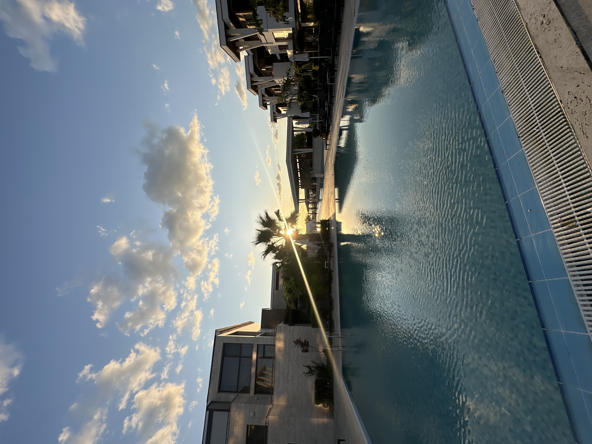 A photograph taken at sunset at a resort in Sozopol, Bulgaria. The image features a large swimming pool in the foreground, with clear blue water reflecting the sky and surrounding buildings. The pool is bordered by a tiled edge and a grated drain.
In the midground, palm trees frame the setting sun, which casts a warm glow and creates lens flare across the scene. Adjacent to the pool are lounge chairs and umbrellas, suggesting a recreational area. Modern hotel buildings with clean lines and large windows are visible on either side of the pool. One building on the left is made of light-colored stone, while buildings on the right feature a more contemporary design with prominent white supports.
The sky is a vibrant blue, dotted with fluffy, golden-tinged clouds illuminated by the setting sun. The overall impression is one of a serene and luxurious holiday destination.