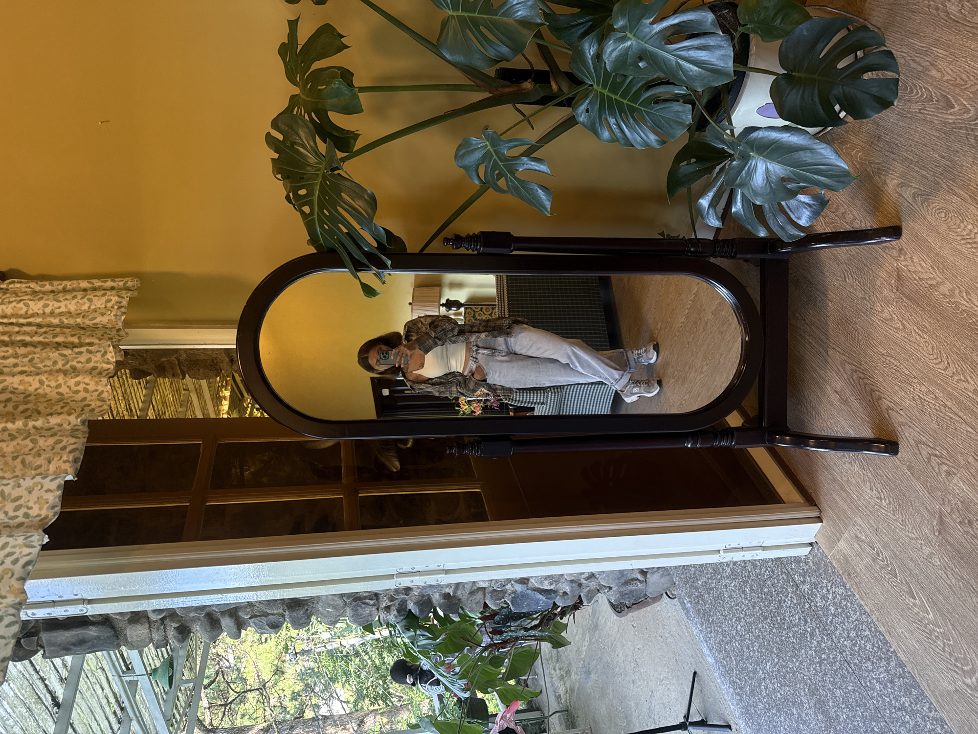 The image depicts an indoor scene with an open doorway leading to an outdoor area. A full-length dark wood mirror stands on a wooden floor, reflecting a young woman taking a selfie. She is wearing a white tank top, a plaid shirt, light-colored pants, and sneakers. The room features a yellow wall, a large Monstera plant in a white pot with purple accents, and a patterned curtain hanging from above. Through the doorway, a stone wall and lush greenery are visible, suggesting an outdoor patio or balcony. The flooring transitions from wooden planks to a speckled gray surface. A tripod and a green pendant lamp can be seen in the outdoor area. The overall ambiance is casual and relaxed. The scene is likely located in Da Lat, Vietnam, known for its cool climate, French colonial architecture, and abundant greenery.