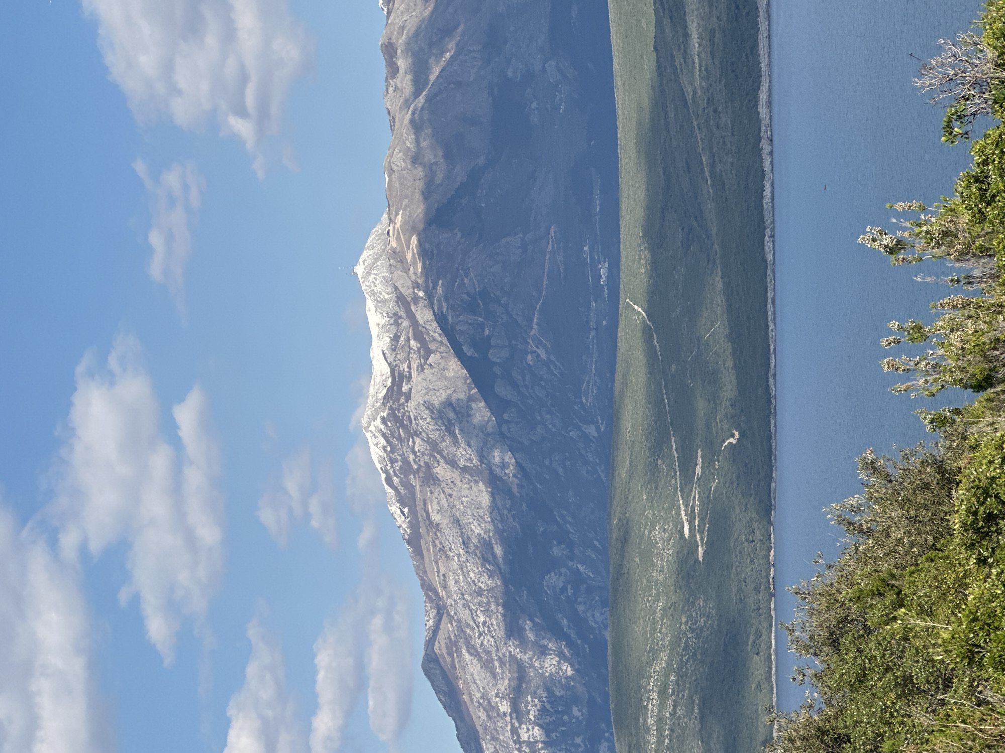 The image captures a landscape near Radovići, Montenegro, featuring a tall mountain with snow-capped peaks on the left side, a body of water on the right, and a green hillside in between. A blue sky dotted with scattered clouds fills the upper portion of the image. Bushes and vegetation appear in the foreground on the lower right side.