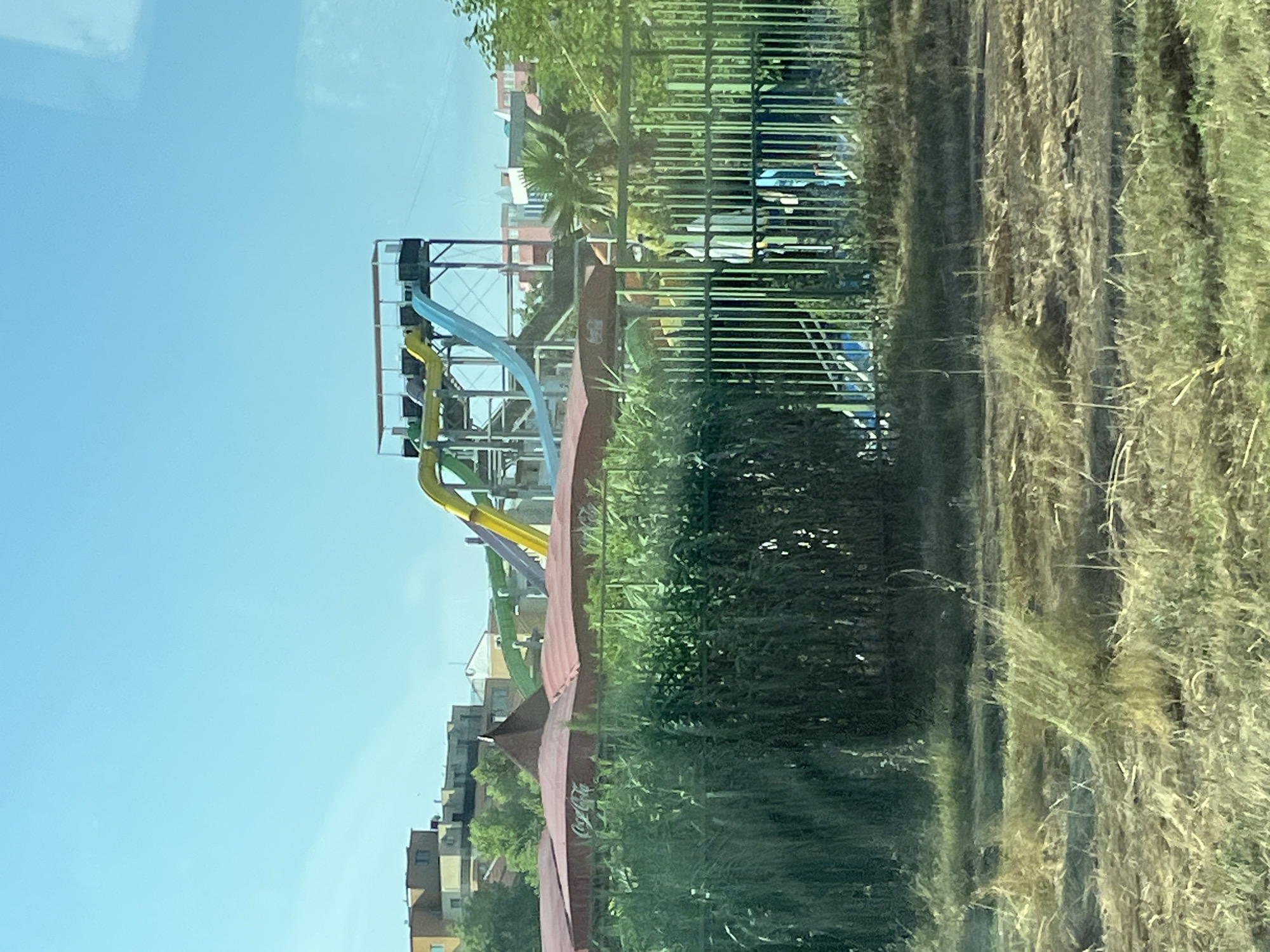 The image depicts a water park in Nesebar, Bulgaria, on a clear day under a bright blue sky. In the foreground, a dense green hedge and a metal fence partially obscure the view. Behind the hedge and fence, a large red awning with the Coca-Cola logo is visible, suggesting a food or seating area within the park. Dominating the midground and background are several colorful water slides, including a yellow, a green, and a blue slide, all connected to a tall platform structure. The slides appear to be made of a slick material suitable for water flow. In the far background, various buildings, some appearing residential or commercial, are visible, interspersed with trees and other foliage. The overall scene suggests a recreational area, likely operational given the presence of the water slides.