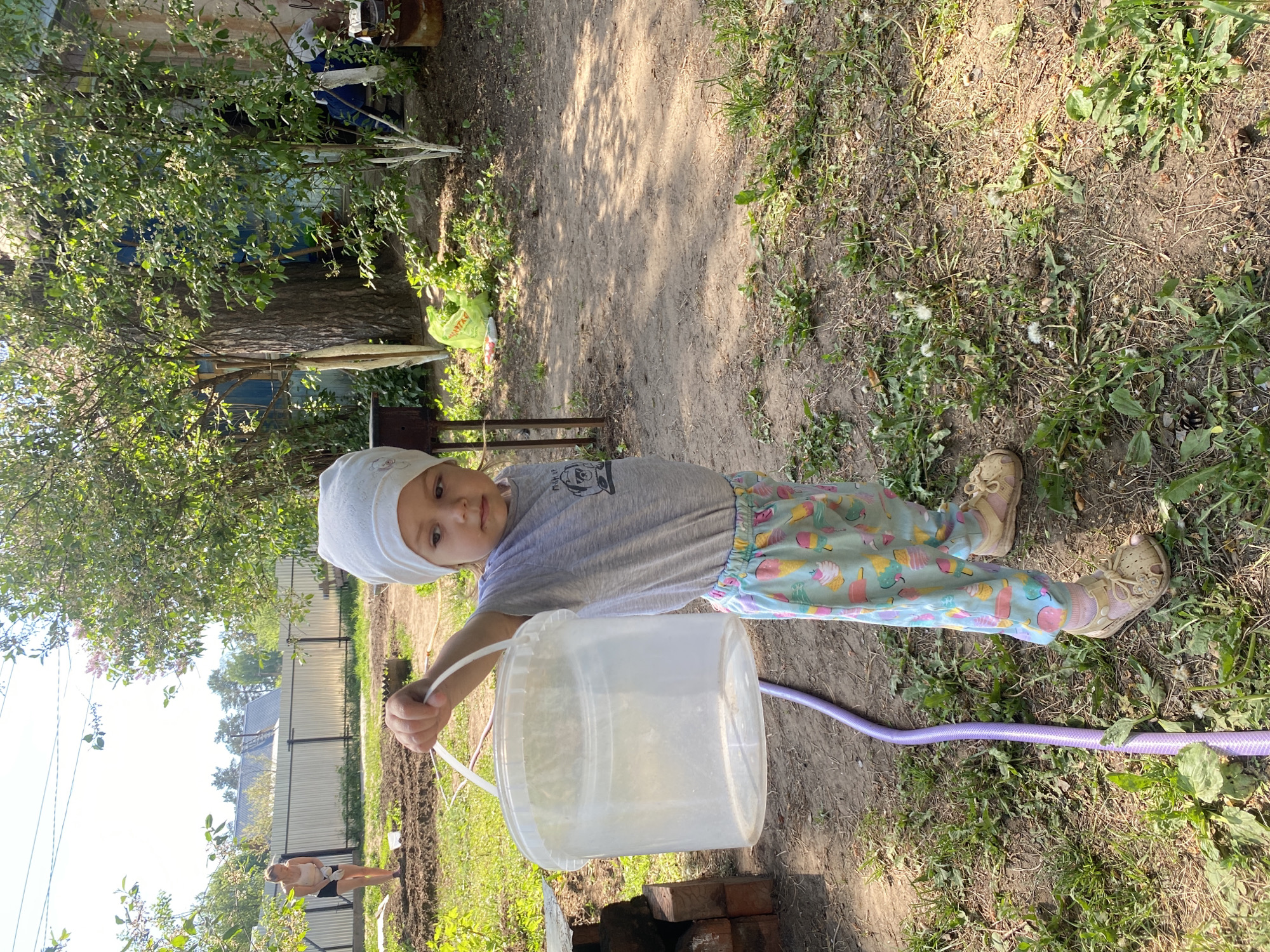 A child in a white headscarf, gray shirt, and patterned pants stands in a grassy area in Chaikovsky, Russia, holding a transparent plastic bucket. A purple garden hose lies on the ground near the child. In the background, there is a dirt path, trees, a fence, and a person in a black swimsuit standing in a garden. Bricks are stacked near the bucket.