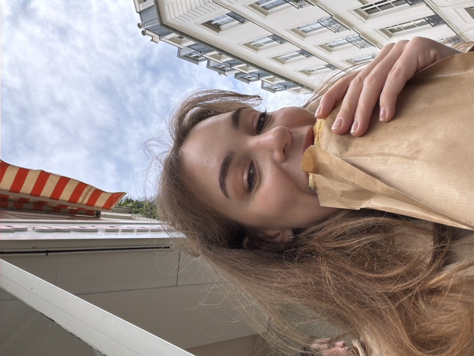 The image depicts a close-up, low-angle selfie of a young adult female on a street in Paris, France. She has long, light brown hair and is smiling, looking directly at the camera with her eyes partially closed. Her right hand holds a food item, possibly a crepe or sandwich, wrapped in a plain brown paper bag, which she is actively biting into. A small silver hoop earring is visible in her left ear. In the blurred background to the lower left, a partial profile of another individual, possibly male, is visible. The background features classic Parisian architecture: a building with a red and white striped awning on the left, and a multi-story, light-colored Haussmannian building with windows and dark wrought-iron railings and a grey mansard roof on the right. A cloudy blue sky is visible above.