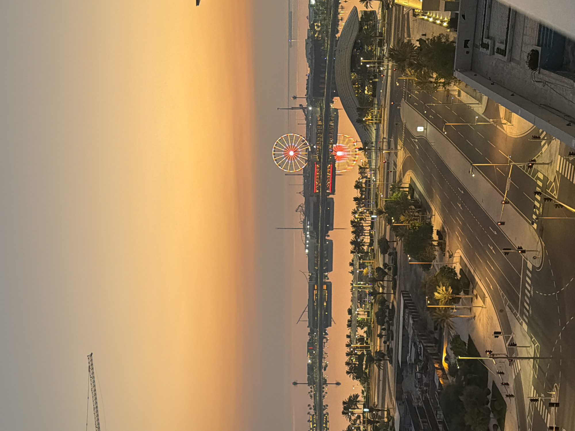 A high-angle, wide shot captures a waterfront city scene at dusk. The sky is a gradient of warm orange and soft gray, transitioning into a hazy horizon. In the distance, a large Ferris wheel illuminated with red and yellow lights dominates the skyline, its reflection shimmering on the calm water. Along the waterfront, various buildings and structures are visible, some with their lights on, casting a warm glow. Palm trees are scattered throughout the landscape, and streetlights line the roads in the foreground. A construction crane extends from the left side of the frame, reaching into the sky. The foreground features a road network with streetlights and traffic signals, indicating an urban environment. The water appears still, creating a mirrored image of the illuminated structures.