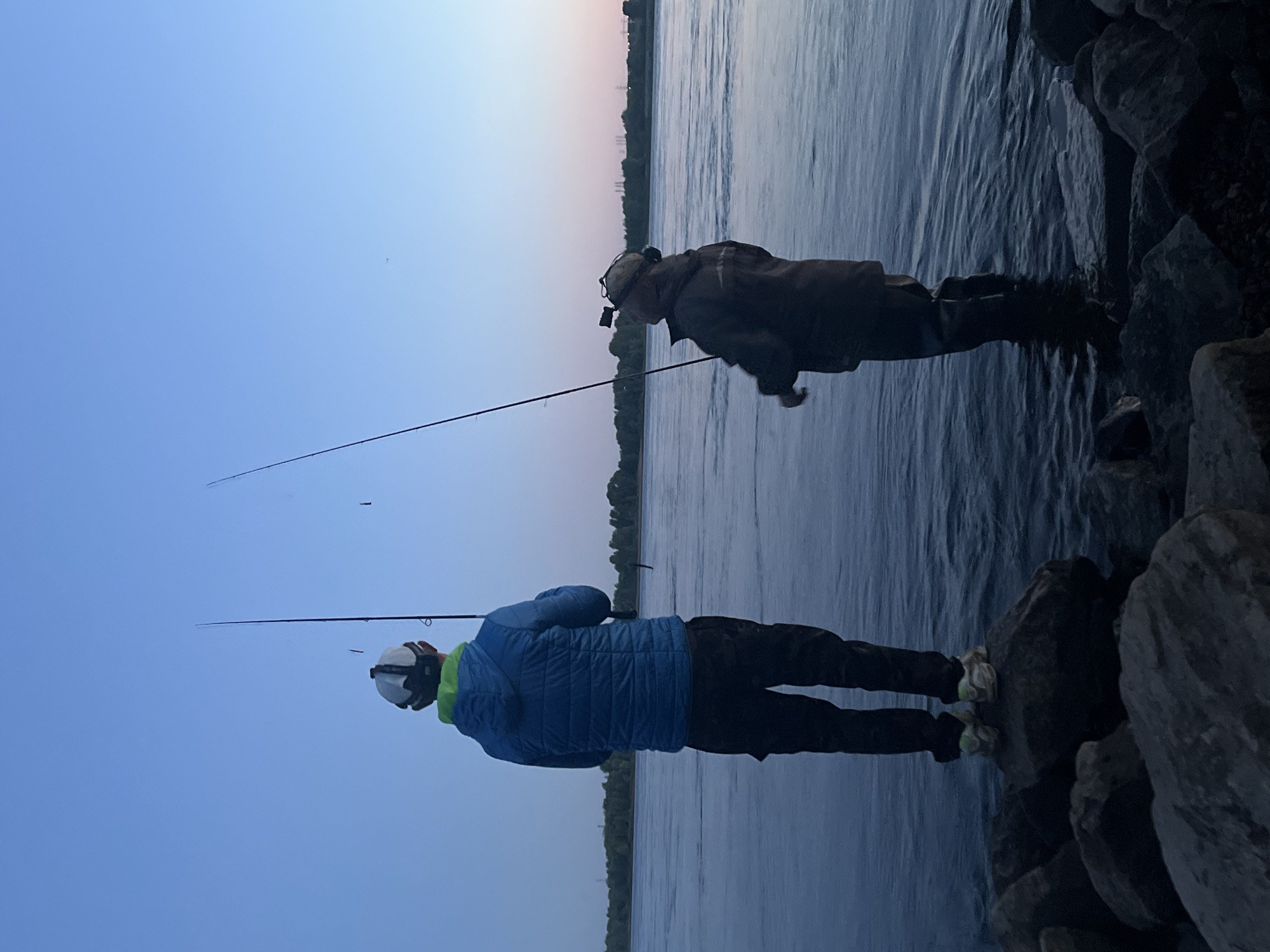 Two individuals are fishing in the Kama River at dusk, near Chaikovsky, Russia. The person on the left, viewed from behind, wears a blue puffer jacket, black pants, and a white and green cap. They are standing on large rocks at the river's edge and holding a fishing rod. The second individual, also viewed from behind and to the right, is partially submerged in the water, wearing a dark jacket, dark pants, and a cap with a head-mounted camera. They are also holding a fishing rod. The river is calm with a visible shoreline in the background lined with trees and structures, under a clear blue sky with a hint of pink near the horizon. Rocks are scattered in the foreground.
