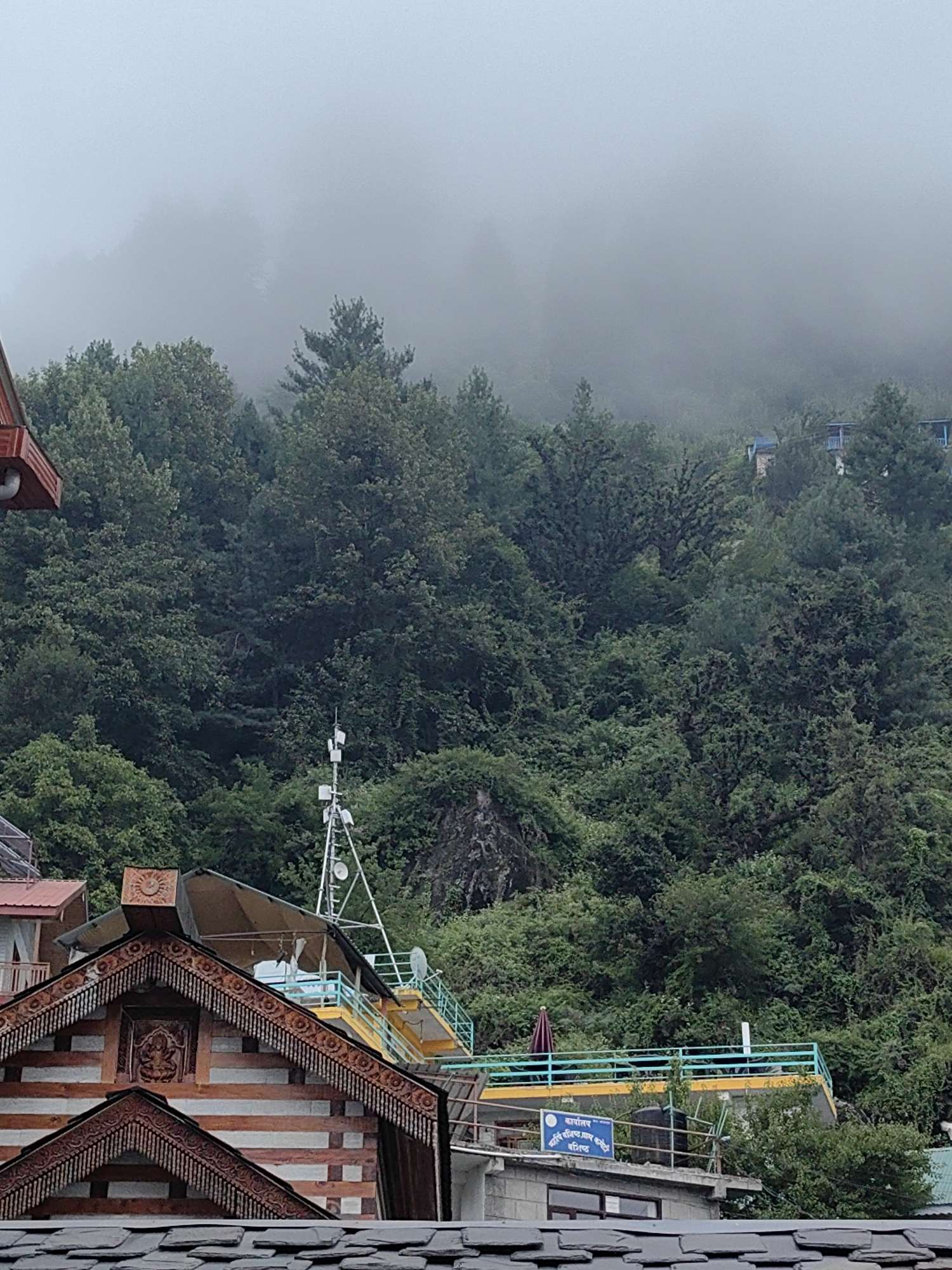 The image depicts a misty mountain scene in Vashisht, India. In the foreground, traditional Himachali architecture with intricate wooden carvings and slate roofs is visible. A multi-story building with a distinctive wooden facade and ornamental eaves is prominent. Behind this structure, a telecommunications tower stands tall, equipped with antennas and satellite dishes. Further up the densely forested mountainside, several buildings with blue railings and yellow accents can be seen nestled amongst the trees, partially obscured by fog. The natural environment is characterized by lush green vegetation, with the mist creating a serene and somewhat ethereal atmosphere. The sky is overcast and grey, indicating cloudy or rainy weather. There are no people visible in the frame.