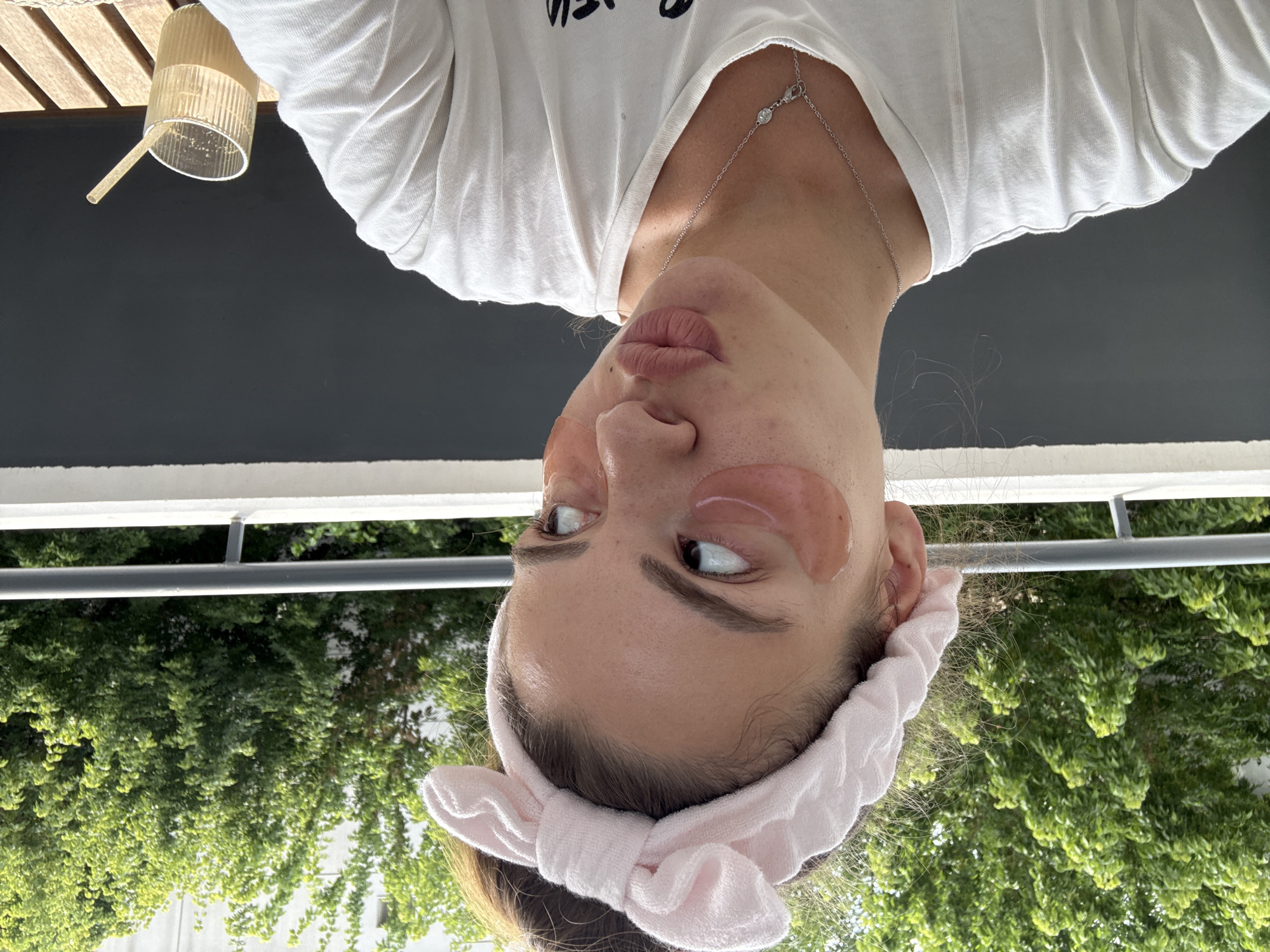 A young woman with a pink headband and pink under-eye masks makes a kissing face at the camera. She is wearing a white t-shirt and a silver necklace. She is standing on a balcony overlooking trees and buildings. A glass with a straw and a drink is on a wooden table to her right. The image appears to have been taken on a sunny day.