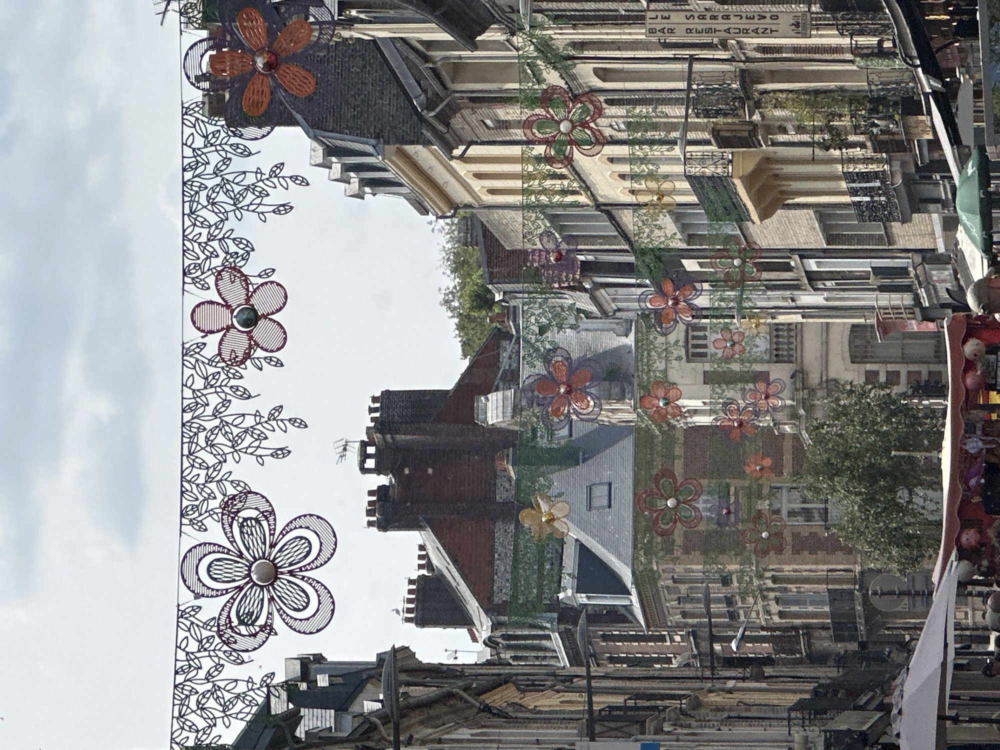 This is a street scene in Dieppe, France. The perspective is looking down a narrow street lined with multi-story buildings, with the sky visible above. Decorative flower-shaped garlands, made of wire and featuring colorful elements, are strung across the street, creating an overhead canopy. These decorations appear to be for a festival or event. The buildings on either side of the street are a mix of architectural styles, with some featuring mansard roofs and brickwork, while others have lighter colored facades. Many buildings have windows with shutters, and some have balconies. In the foreground, parts of market stalls or awnings are visible, suggesting a market is taking place or has recently concluded. There are a few people visible in the lower part of the frame, some appearing to be shoppers or vendors. The overall impression is a lively urban street during daylight hours, decorated for a festive occasion.