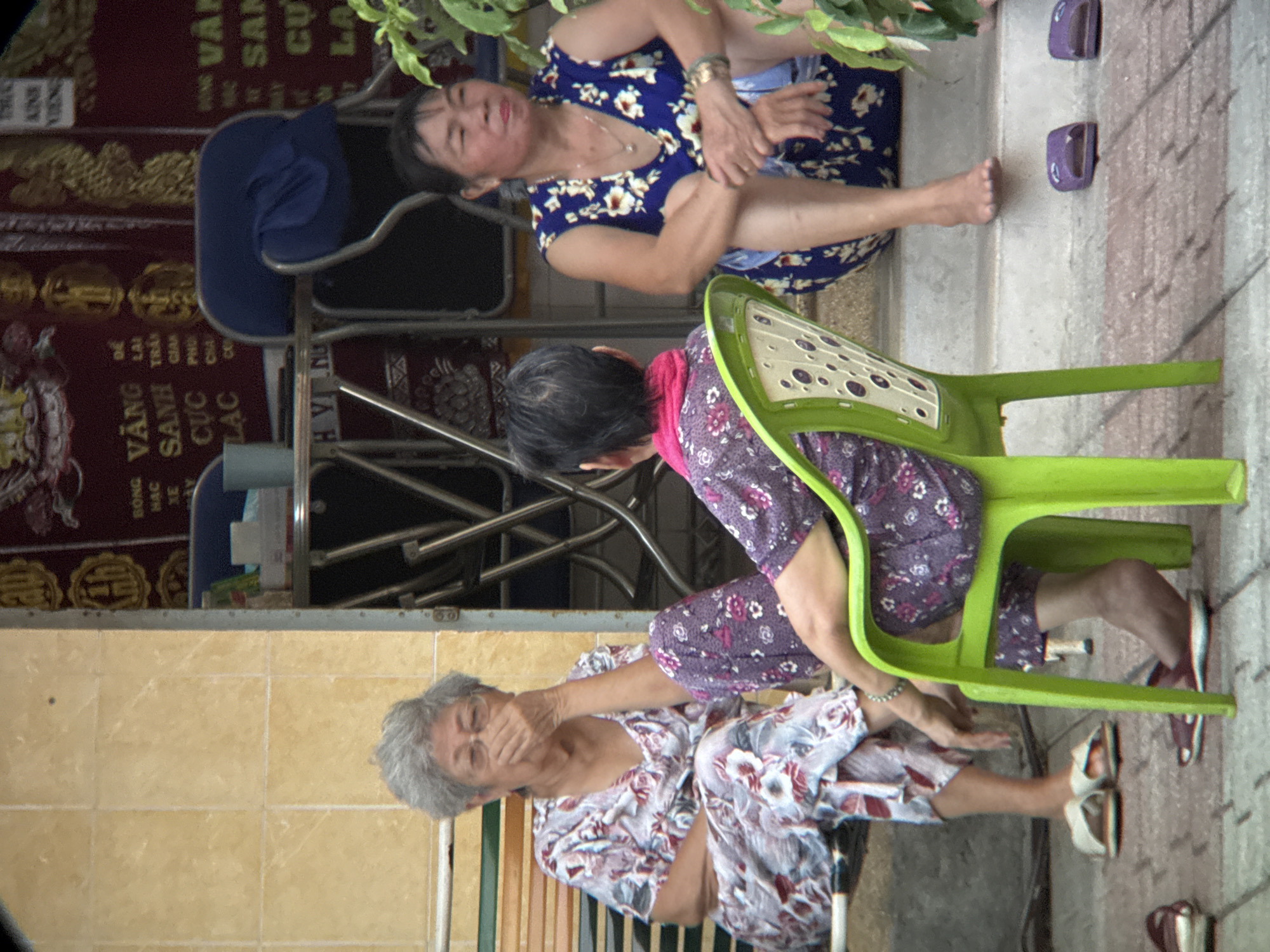 The image depicts a street scene in Nha Trang, Vietnam, with three women of Asian ethnicity seated in front of a building. The woman on the left, wearing a floral patterned pajama set and white sandals, is seated on a bench and is covering her mouth with her hand. To her right, another woman in a similar floral pajama set is partially obscured by a bright green plastic chair, on which she is seated. She is facing away from the camera. The woman on the far right, dressed in a blue floral sleeveless dress and wearing an ankle bracelet, is sitting on a concrete step, with her legs crossed and feet bare. She is looking towards the left. The background features a building with yellow tiled walls, a red banner with Vietnamese writing, and metal chairs and tables. Green foliage is visible on the right side of the frame. The ground is paved with bricks. The overall impression is one of a casual, outdoor gathering.