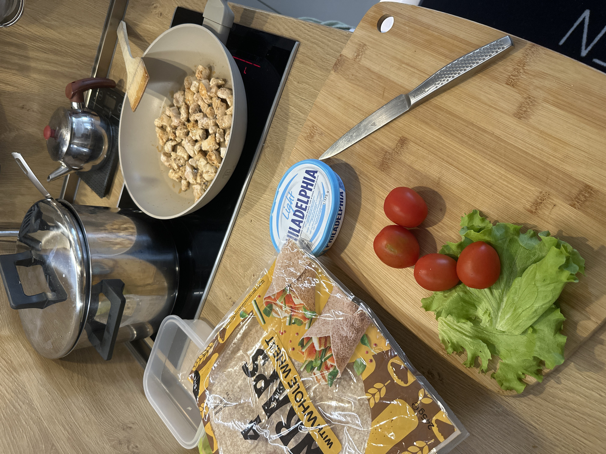 The image depicts a culinary preparation scene on a wooden countertop. A light-colored frying pan on a black stovetop contains diced, cooked chicken pieces. Beside it, a wooden cutting board holds a silver knife, a blue container of Philadelphia cream cheese, three red cherry tomatoes, and a handful of green lettuce. A package of "Bess with Whole Wheat" wraps lies on the counter, partially obscuring a clear plastic container. A stainless steel teapot and a pot with a lid are also visible near the stove. The scene suggests someone is preparing a wrap or sandwich with the ingredients present. There is no information that would place this scene in the city of Plovdiv, Bulgaria.