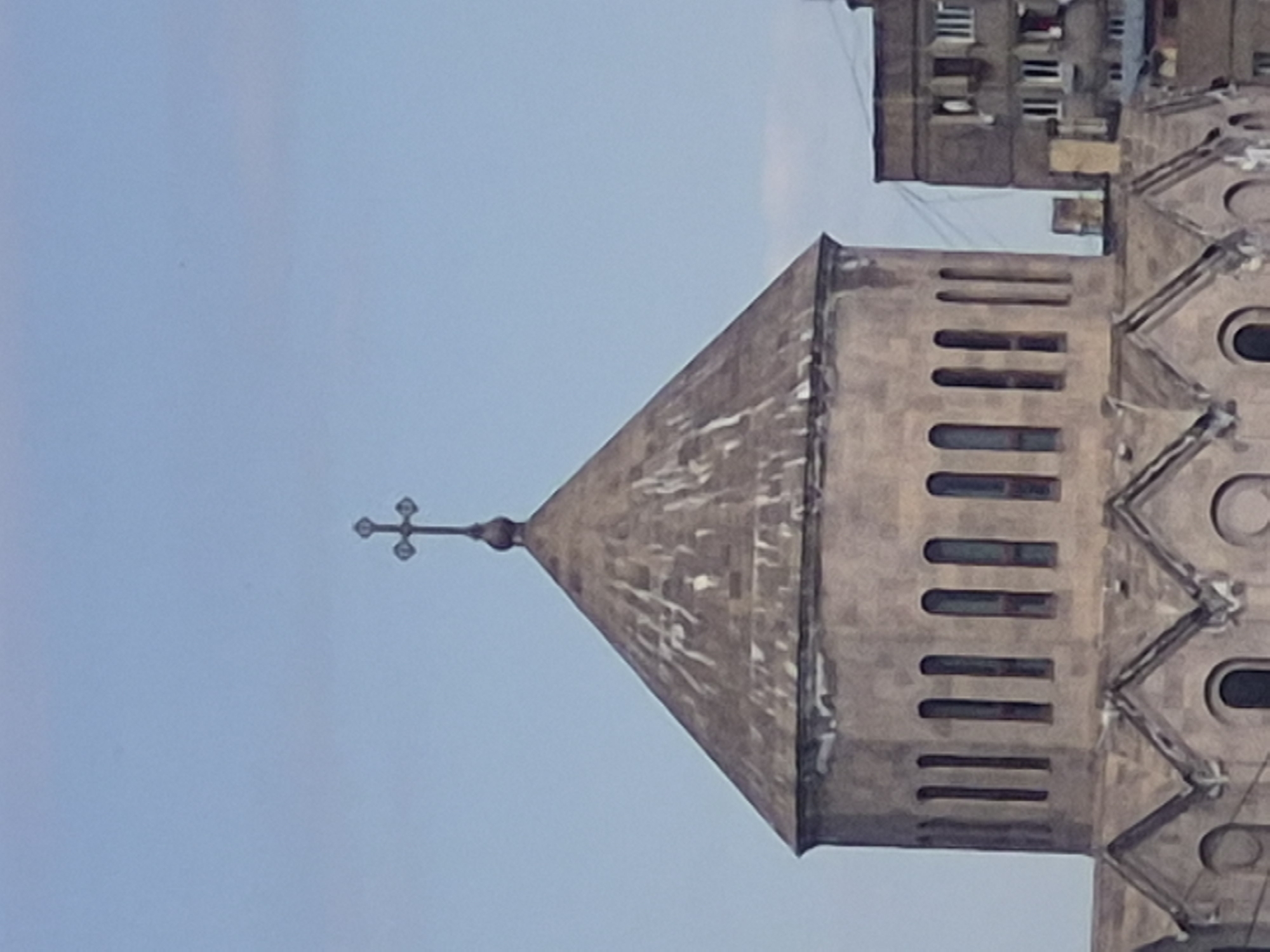 The image displays the upper section of a stone church or cathedral tower against a clear, light blue sky. The tower is constructed from light-colored stone, possibly tufa, and culminates in a prominent, pointed spire. Perched atop the spire is an ornate cross, featuring small decorative elements at the ends of its arms, indicative of an Armenian-style cross.
Below the spire, the tower's belfry or upper section showcases several vertically elongated, rounded-arch openings arranged in vertical rows. The visible portion of the church's main body exhibits intricate architectural details, including a distinctive zig-zag or chevron-like relief pattern on its facade.
In the background, a multi-story residential building is partially visible. This structure, appearing to be made of darker concrete or stone, features numerous rectangular windows and balconies, typical of apartment blocks found in urban settings.
The scene is set during the daytime, likely late morning or afternoon, characterized by soft, diffused lighting and the absence of harsh shadows. The sky is clear, without any visible clouds, suggesting fair weather. There is no discernible activity or any people in the frame. The architectural style of the church and the presence of the residential building are consistent with the provided location context of Yerevan, Armenia. No text is visible in the media file.