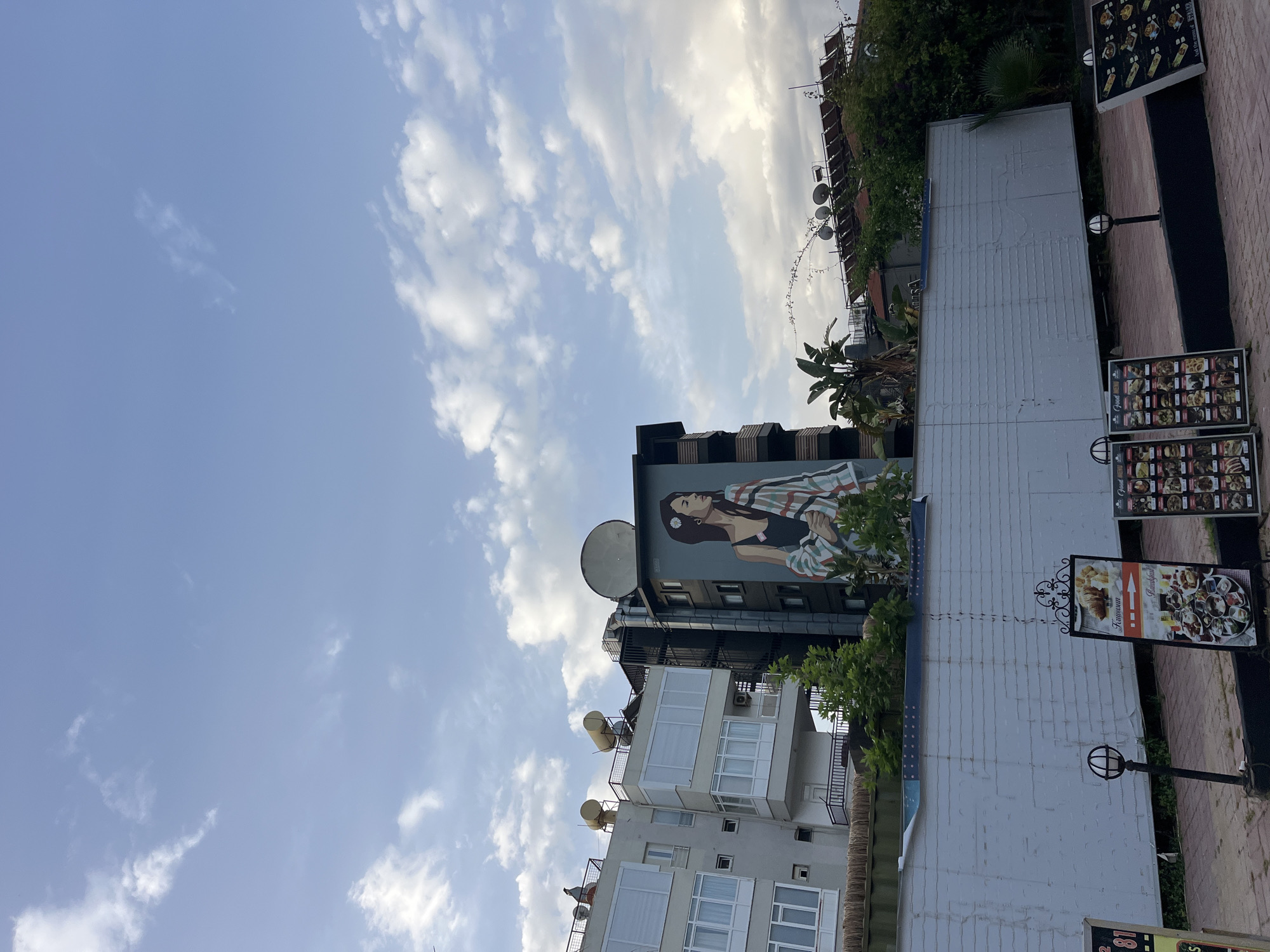 The image shows a street scene in Alanya, Turkey. In the background, there are several buildings with satellite dishes on their roofs. One of the buildings has a large mural of a woman on its side. In the foreground, there is a light-colored building with menu boards displayed outside. The sky is light blue with fluffy white clouds.