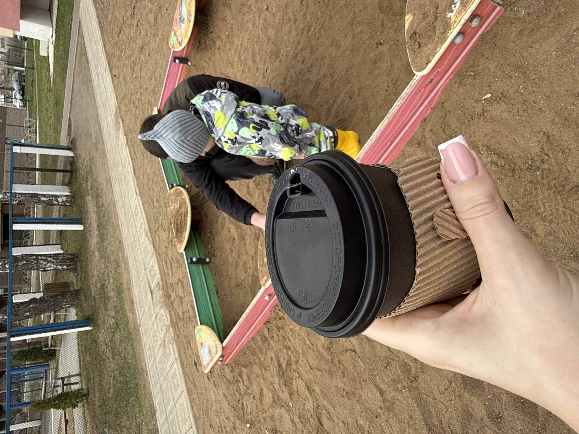 Observed is an eye-level outdoor scene, likely captured in the city of Chaikovsky, Russia. A child wearing a grey hat, black jacket, patterned outerwear and yellow boots, is situated on a playground seesaw, interacting with the equipment. A hand holds a coffee cup with a black lid and a brown corrugated sleeve. The playground is situated on a sand-covered ground with a sidewalk along the left side. In the background are glimpses of buildings, trees, and a blue and white fence.