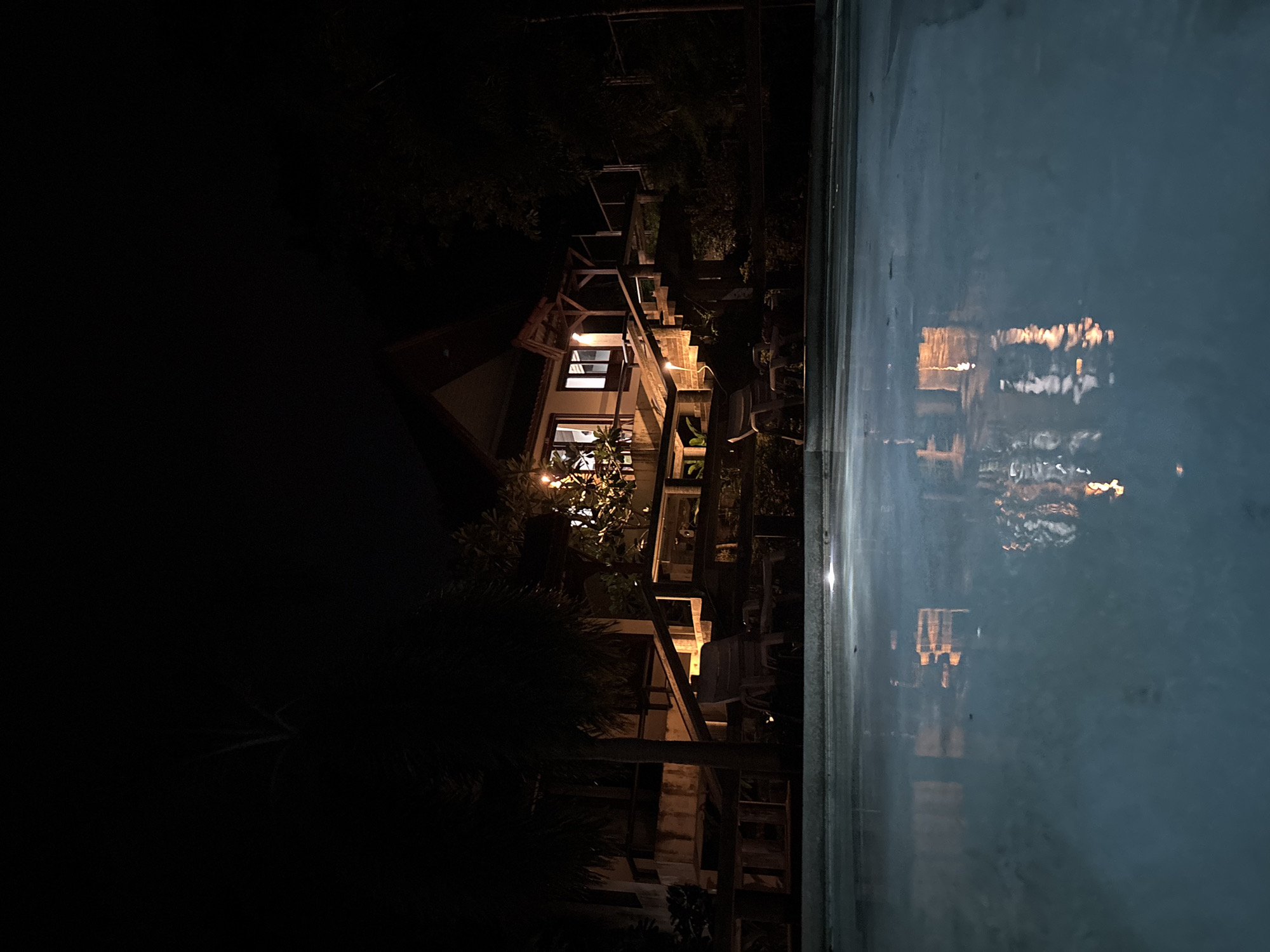 This nighttime image, captured in บ้านบางรักษ์, Thailand, features an illuminated elevated building and a swimming pool. The building, characterized by a peaked roof, presents multiple windows emitting warm yellow light. A multi-tiered wooden deck with railings and a staircase ascends towards the building's entrance. To the right of the main structure, a smaller, illuminated covered porch is visible. The foreground is occupied by a calm, dark blue swimming pool that reflects the building's lights. Two light-colored plastic lounge chairs are situated on a paved surface near the left edge of the pool, partially obscured by a large palm tree. Dense, dark tropical vegetation, including additional palm trees, surrounds the property, with some foliage near the building illuminated by ambient light. No individuals are present in the scene.