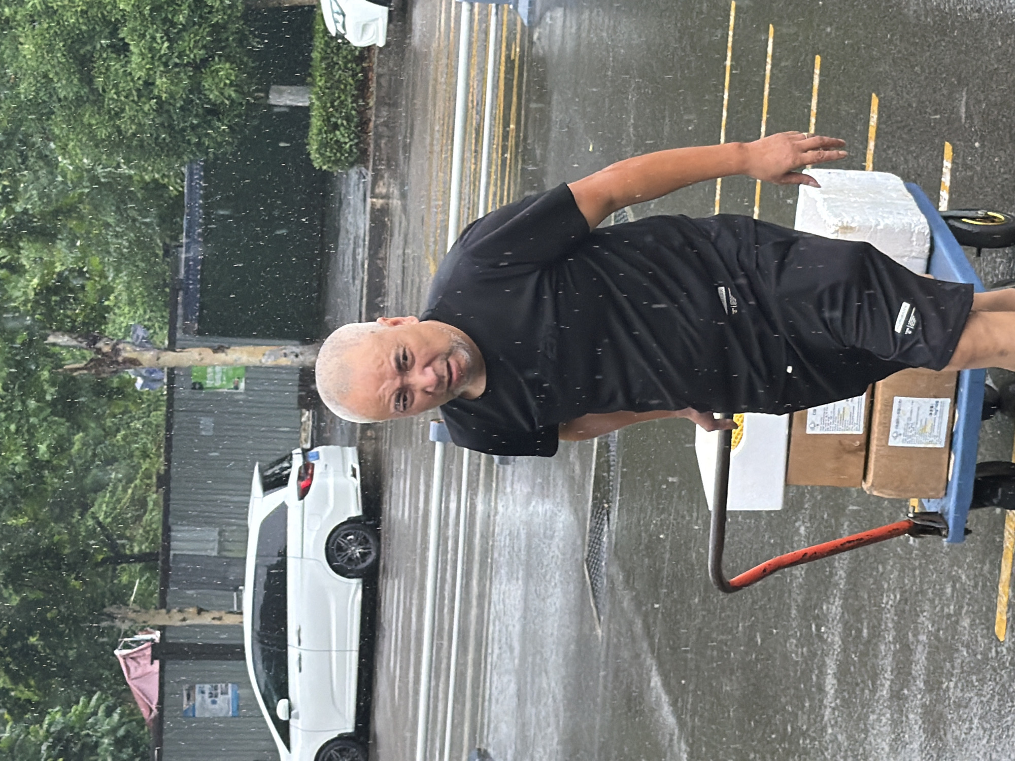 The image displays an urban street scene under rainfall, consistent with a city environment in China, such as Jiangjin. In the foreground, a single adult male with a bald head and light facial stubble walks from left to right across a wet asphalt road. He wears a black short-sleeved t-shirt and black shorts, both featuring white labels. His gaze is directed towards the viewer with an intense expression. He is pushing a blue hand truck with a red handle, which carries a stack of three items: a white, possibly styrofoam, box containing yellow material on top, followed by two brown cardboard boxes underneath, one with a visible white label containing Chinese text.
The street surface is wet, reflecting active precipitation, with numerous raindrops visible against the background. Yellow dashed lines are painted on the asphalt. A metal drainage grate is embedded in the road. Behind the man, a white Honda Odyssey minivan with dark aftermarket wheels and tinted windows is parked on the left, partially obscured by a metal barrier featuring white and yellow stripes. A partial blue license plate is visible on the minivan's rear. The front section of another white vehicle is visible on the far right. The background features a corrugated metal fence, likely construction hoarding, bearing blue and green signs with Chinese characters. Green trees and dense foliage are visible above and behind the fence, with a tree trunk prominent on the far left.
