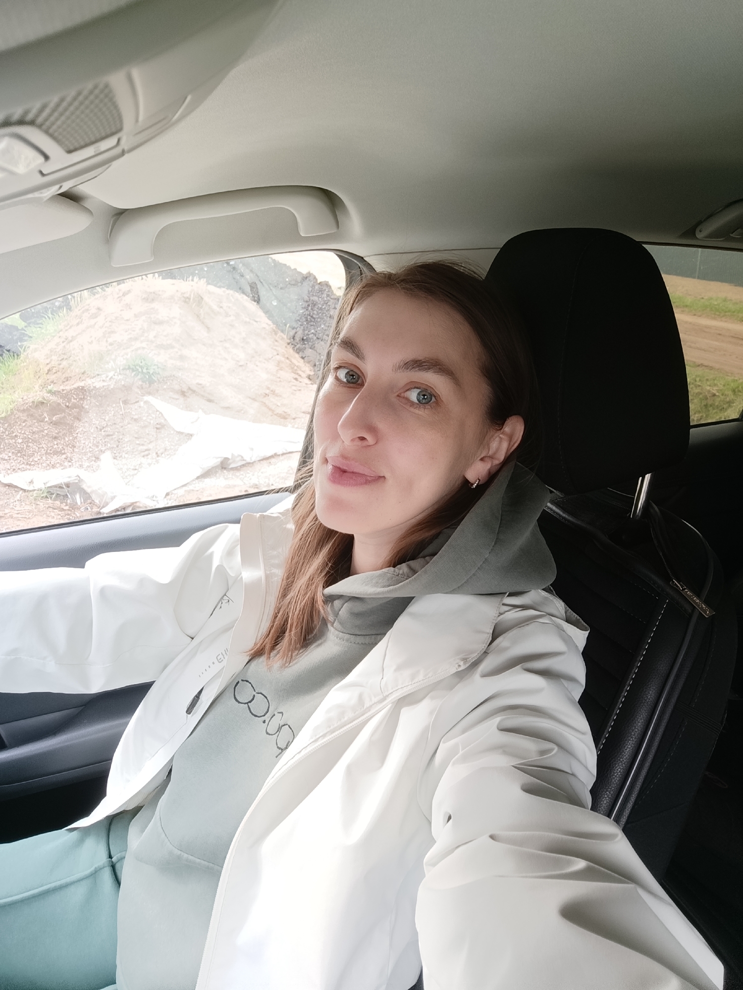 A woman is taking a selfie inside a car. She has light skin, blue eyes, and light brown hair. She is wearing a gray hoodie and a white jacket. Through the car window, there is a view of a dirt pile and some vegetation. It is possible that this photograph was taken in Chaikovsky, Russia, based on the user's request. The car's interior is visible, with black leather seats. The seat headrest is black. The car's ceiling and handles are light gray. The background shows a dirt road and green vegetation.