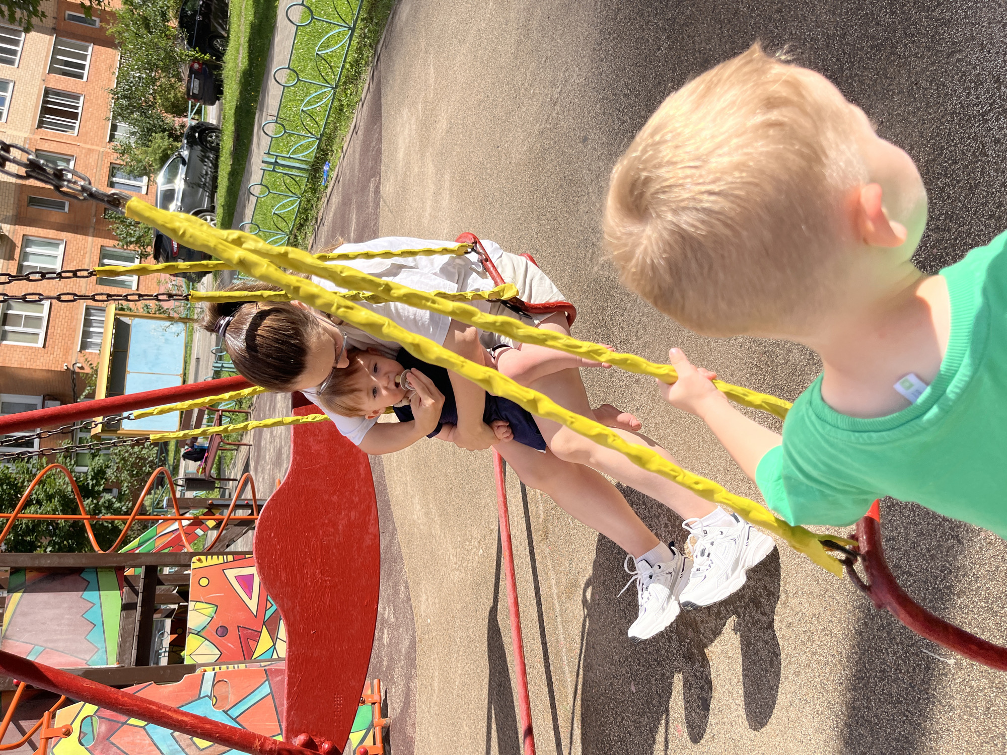 In Balashikha, Russia, on a sunny day, a mother and her two young children are at a playground. The mother, seated on a red swing, holds a baby in her arms. The baby is wearing a dark blue outfit and has a pacifier in its mouth. The mother is dressed in a white t-shirt and light-colored shorts, with white sneakers on her feet. In the foreground, the back of another child's head is visible, wearing a bright green t-shirt. This child appears to be reaching out towards the swing. The playground features a colorful structure with geometric patterns. In the background, a brick apartment building and parked cars are visible, along with a grassy area bordered by a decorative metal fence. The ground of the playground is a textured surface.