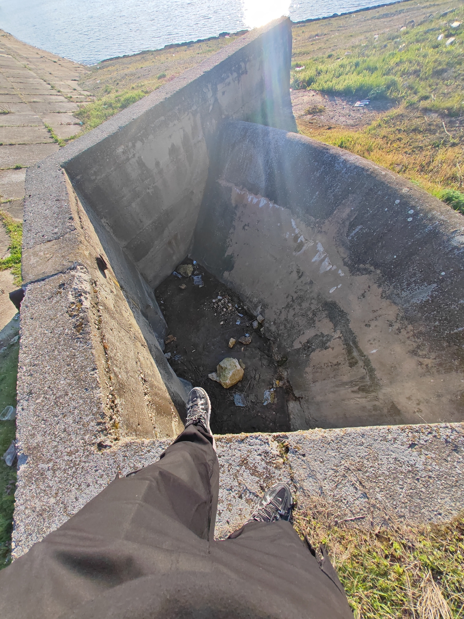 A person stands on the edge of a large, concrete structure resembling a drainage channel or a type of water management system. The person is wearing black pants and athletic shoes with grey and black patterns. The structure is filled with debris, including rocks, trash, and standing water. The location appears to be near a body of water, visible in the background. The surrounding area is grassy and unkempt, with patches of vegetation. The sunlight suggests it is daytime. The image was taken in Shumen, Bulgaria.