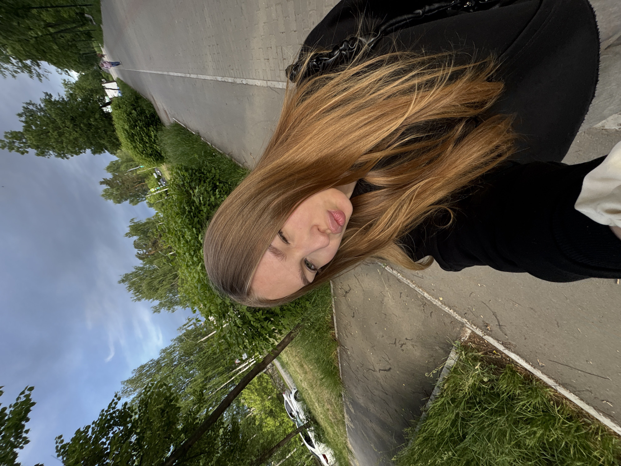 A young woman with long blonde hair poses for a selfie on a path. She is wearing a black shirt or jacket. In the background, there is a tree-lined path and a cloudy sky. White cars are parked along the path. The location is a city park path in Chaikovsky, Russia.