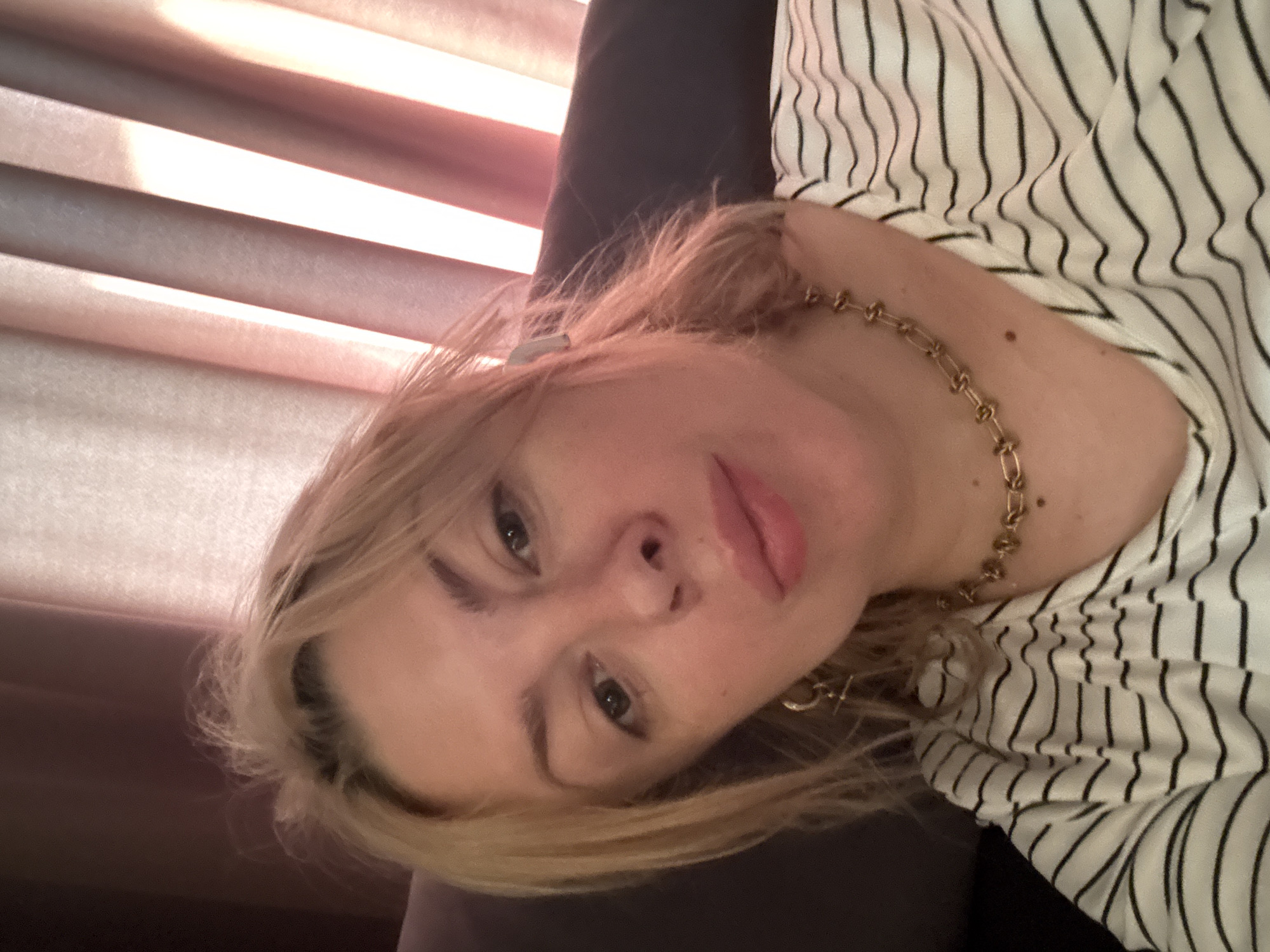 A light-skinned woman with blonde hair is taking a selfie. She is wearing a striped shirt and a gold necklace. The background appears to be a room with curtains. The lighting suggests it might be daytime. There is no indication of the location being in Sochi, Russia.