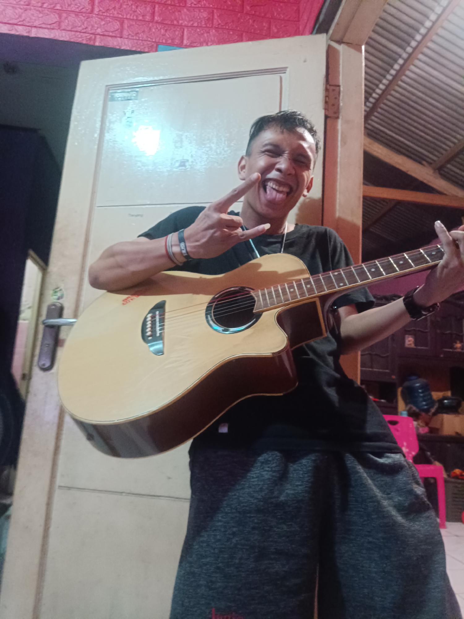 A young man stands indoors, holding an acoustic guitar. He is wearing a black t-shirt and dark grey sweatpants. His mouth is open, and his tongue is sticking out, with a peace sign gesture in his left hand. The man's right arm is extended, holding the guitar neck. The guitar is a light wood color with a darker brown trim around its body. He has bracelets on both wrists. The background shows a cream-colored door and a red brick-patterned wall. A ceiling with exposed beams is visible in the upper right corner. The lighting suggests it is nighttime or an interior space. The overall scene appears to be a casual, personal setting, potentially a home in Depok, Indonesia.