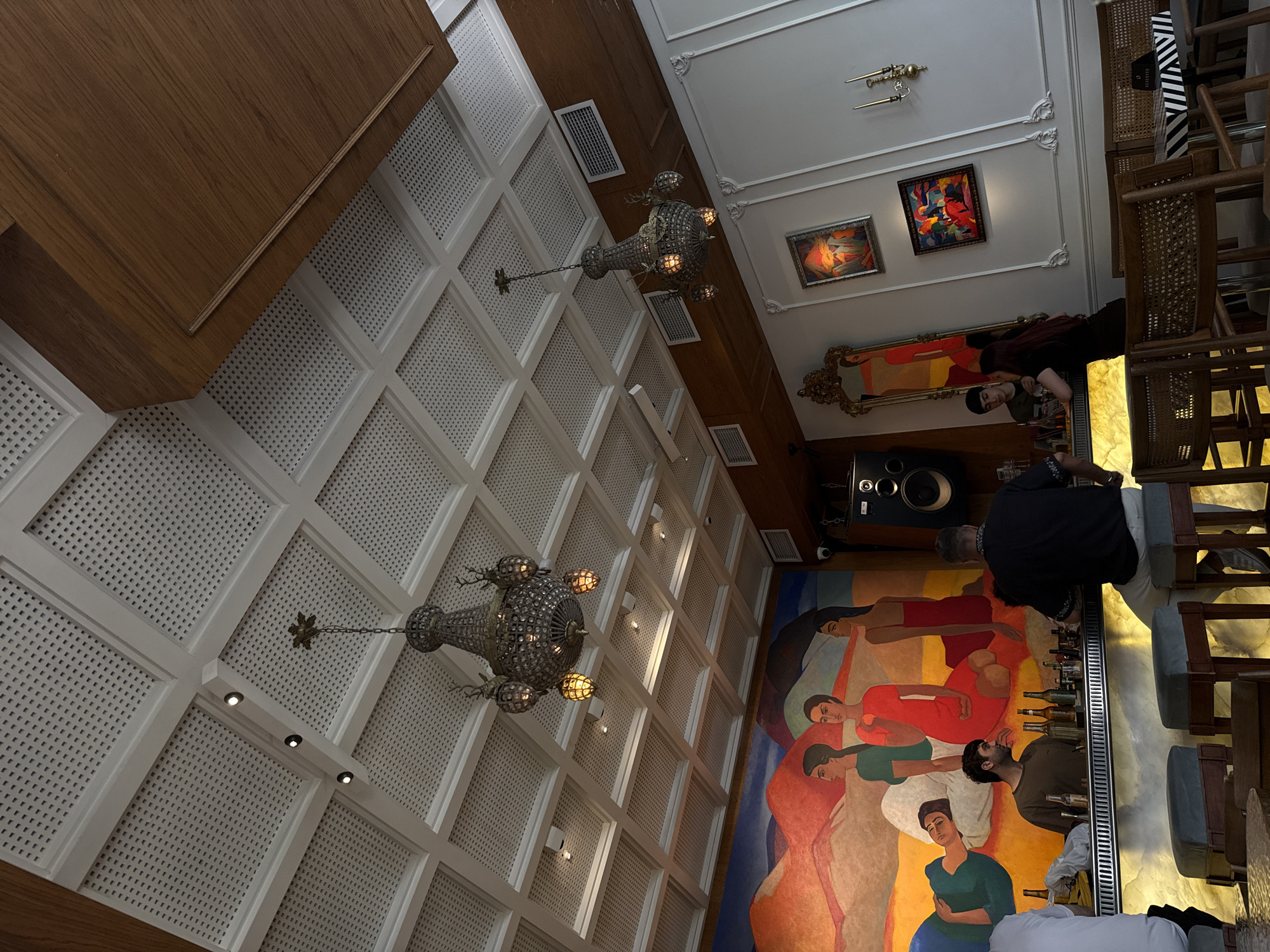 The media file depicts the interior of a modern establishment, likely a bar or restaurant, with a distinctive contemporary design. The scene is characterized by a high, ornate ceiling with a grid pattern and intricate decorative moldings. Two elaborate chandeliers, crafted from crystal or glass and metal, hang from the ceiling, casting a warm glow. Several framed artworks adorn the walls, one particularly vibrant painting featuring abstract figures, and another depicting a more figurative scene with warm tones.
In the foreground, a large, colorful mural covers a significant portion of the wall, showcasing stylized human figures in various poses and vibrant hues. The mural suggests a lively atmosphere and artistic expression.
Several individuals are visible in the scene. One person, dressed in dark clothing, appears to be working behind a bar, arranging bottles. Another person, wearing a dark shirt and a beanie, is seated at the bar, looking towards the camera. Further down the bar, another individual in a black shirt is interacting with someone. The presence of a bar stocked with bottles indicates that drinks are being served.
Objects in the scene include a large speaker system mounted on the wall, various bottles on the bar, and chairs with wooden frames and cushioned seats. The lighting is ambient, with the chandeliers and spotlights creating a warm and inviting atmosphere. The overall impression is of an upscale and artistic venue. The images are captured from a high vantage point looking downwards, suggesting the photographer was on an upper level or on a ladder. The presence of art and the design elements suggest the location could be a trendy spot in Yerevan, Armenia.