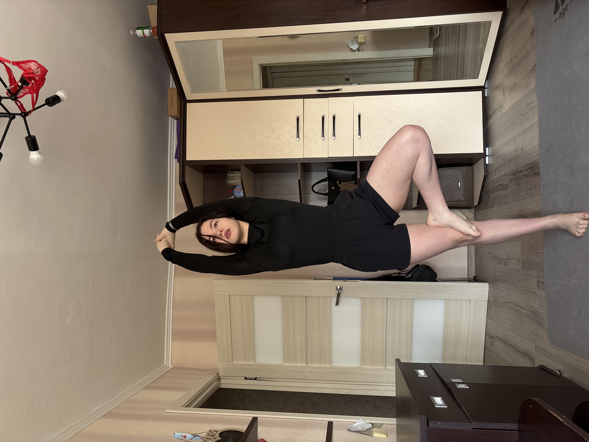 A young female, barefoot, stands on a grey yoga mat in a residential room in Perm, Russia. She is performing a balance pose, supporting herself on her left leg with her right foot placed against her left inner thigh. Her arms are raised above her head, hands clasped. She wears a black long-sleeved top and black shorts. A small, dark tattoo is visible on her right ankle.
The room features light-colored vertically striped wallpaper and a white ceiling with crown molding. A black four-arm chandelier with exposed light bulbs hangs from the ceiling; a red lace thong is draped over one of its arms. The floor is covered with light brown laminate wood.
On the left, a partial view of a dark brown cabinet or dresser with silver handles is visible, along with a wall-mounted shelf displaying decorative items. In the center-left background, a light-colored wooden door with frosted glass panels is ajar, revealing a darker space beyond.
On the right side of the room, a large two-tone wardrobe unit with light beige, patterned panels and dark brown framing is present. The wardrobe includes drawers with dark handles and open shelves, one holding a black handbag and a beige pouch. The far-right section of the wardrobe is a full-length mirror, reflecting a view of an adjacent room or hallway that includes a closed door and a weighing scale on the floor. On top of the wardrobe, a bottle and a cardboard box labeled "Open The Future" are placed.