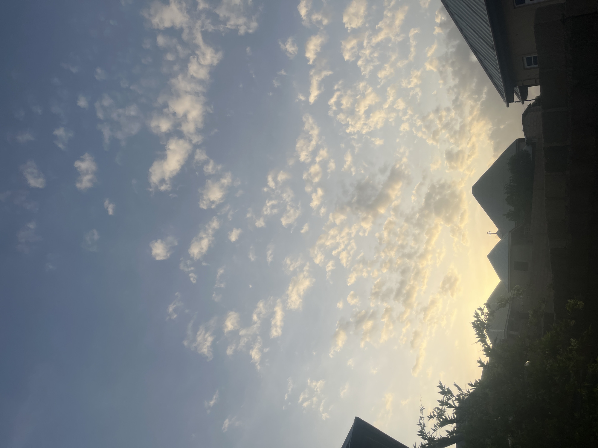 The image shows a silhouetted view of buildings and trees against a sky filled with scattered clouds at dawn or dusk. The sky is a gradient of soft blues at the top, transitioning to pale yellow and orange near the horizon where the sun is not directly visible but its light illuminates the clouds. Several buildings with peaked roofs are visible, some appearing to be houses or residential structures. In the foreground, on the left, is the dark silhouette of dense foliage and a partial view of a building roof. The overall scene is peaceful and suggests an outdoor setting in a semi-urban or rural area, possibly Barakin Akawo, Nigeria, given the architectural style of the roofs and the type of vegetation. No people or animals are discernible in the image.