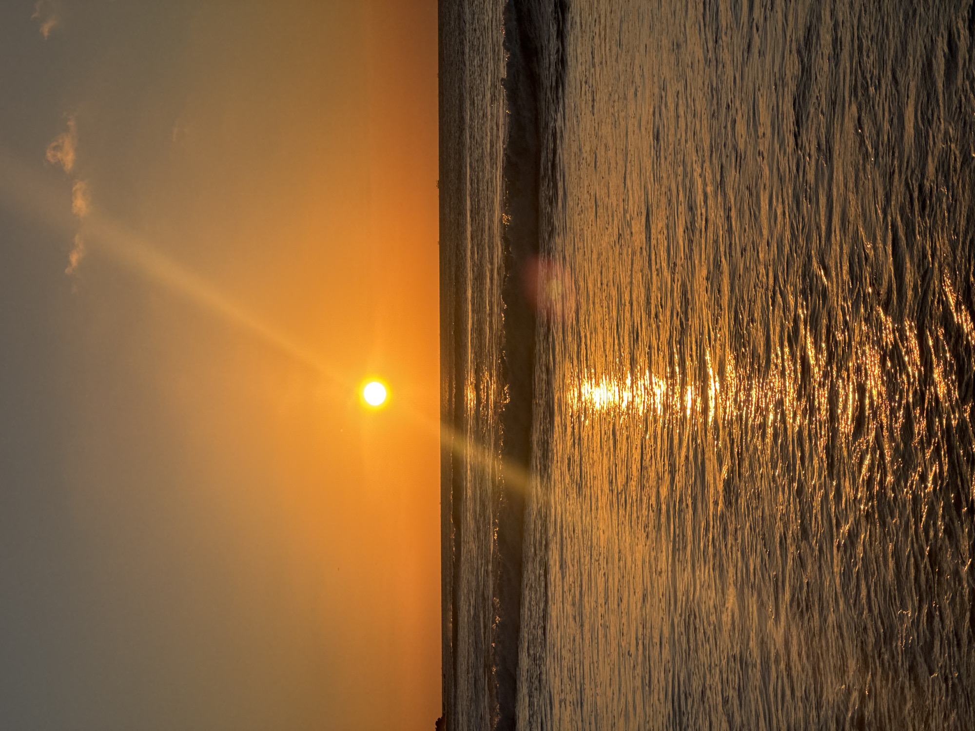 This image displays a serene sunset or sunrise over an ocean, likely in Kuta Selatan, Indonesia. The sun, a brilliant white-yellow orb, is positioned low on the left side of the frame, radiating intense light rays that cut across the sky. The sky is painted with warm hues, transitioning from vibrant orange and yellow closest to the sun to a softer, muted grey-blue in the upper left. A few faint, wispy clouds are visible in the less illuminated parts of the sky.
The ocean dominates the right half of the image, reflecting the sun's golden light with a dazzling, shimmering effect. The water's surface is covered in small ripples or gentle waves, which catch the sunlight to create a textured pattern of sparkling gold and orange streaks. A faint, dark horizon line separates the sky and the water, with a barely discernible strip of land or distant coastline along it.
The scene evokes a sense of tranquility and natural beauty, characteristic of a peaceful coastal morning or evening. The time of day is clearly either sunrise or sunset, indicated by the low position of the sun and the warm, intense lighting. No people or man-made objects are visible, allowing the natural spectacle to be the sole focus. There is no visible text in the media file.