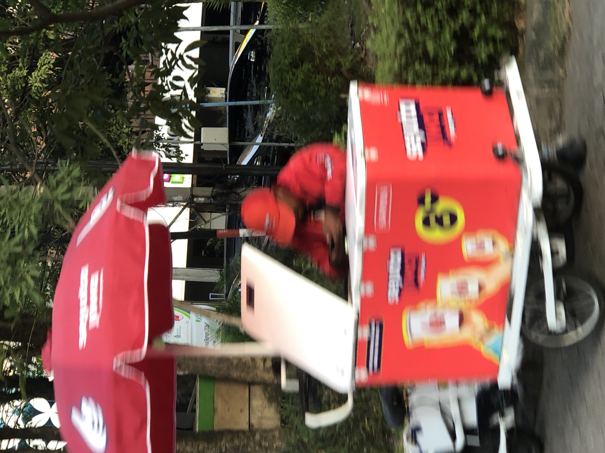 A person wearing a red cap and jacket is seated behind a red cooler cart with a white lid. The cooler cart has branding on its side featuring text and images of cups. The cart appears to be outdoors, with trees and bushes in the background. A red umbrella with white trim is positioned above the cart. The scene suggests a vendor selling items from the cart. The location is identified as Sidoarjo, Indonesia.