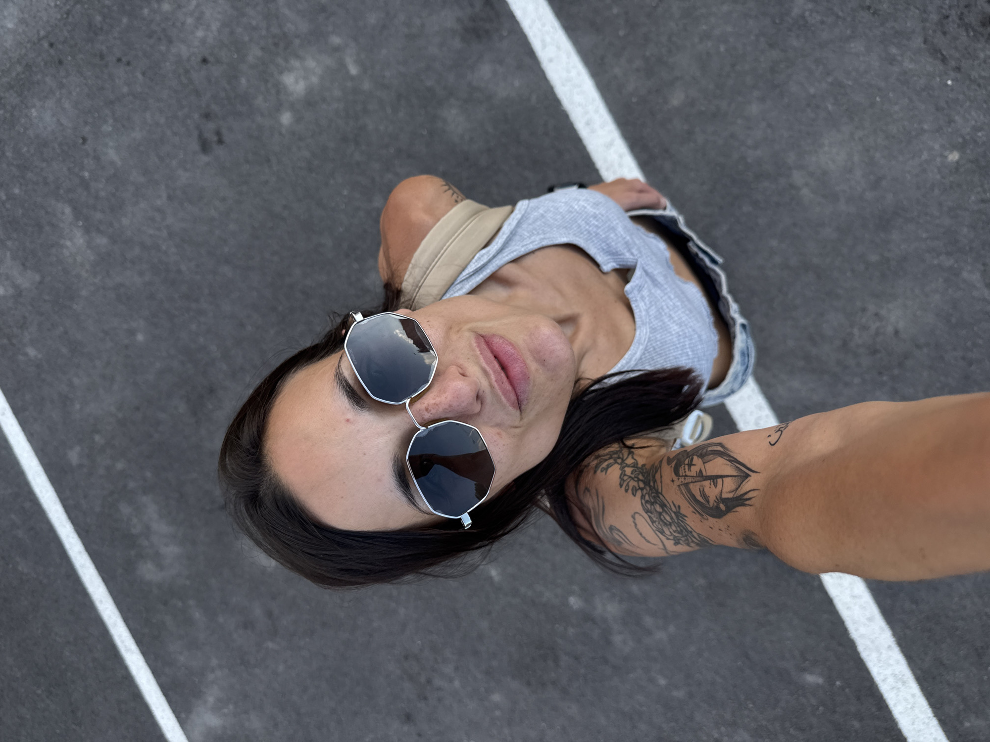 An overhead selfie captured in Pleven, Bulgaria, depicts a woman with dark hair, sunglasses, and multiple tattoos, looking directly up at the camera. Her face shows a slight smile with lips slightly parted, and freckles are visible on her nose and cheeks. She wears silver-framed, dark-lensed, octagonal sunglasses, which reflect a blurred image of the sky. She is dressed in a light grey ribbed crop tank top and light blue denim bottoms. Her left arm is extended towards the camera, prominently displaying several black ink tattoos, including a split-face design on her inner forearm and a floral/vine pattern higher up on her arm. The background consists of dark grey asphalt pavement with two distinct white painted lines: one extending diagonally from the upper left to the middle right, and another straight line positioned horizontally in the lower right section of the frame.