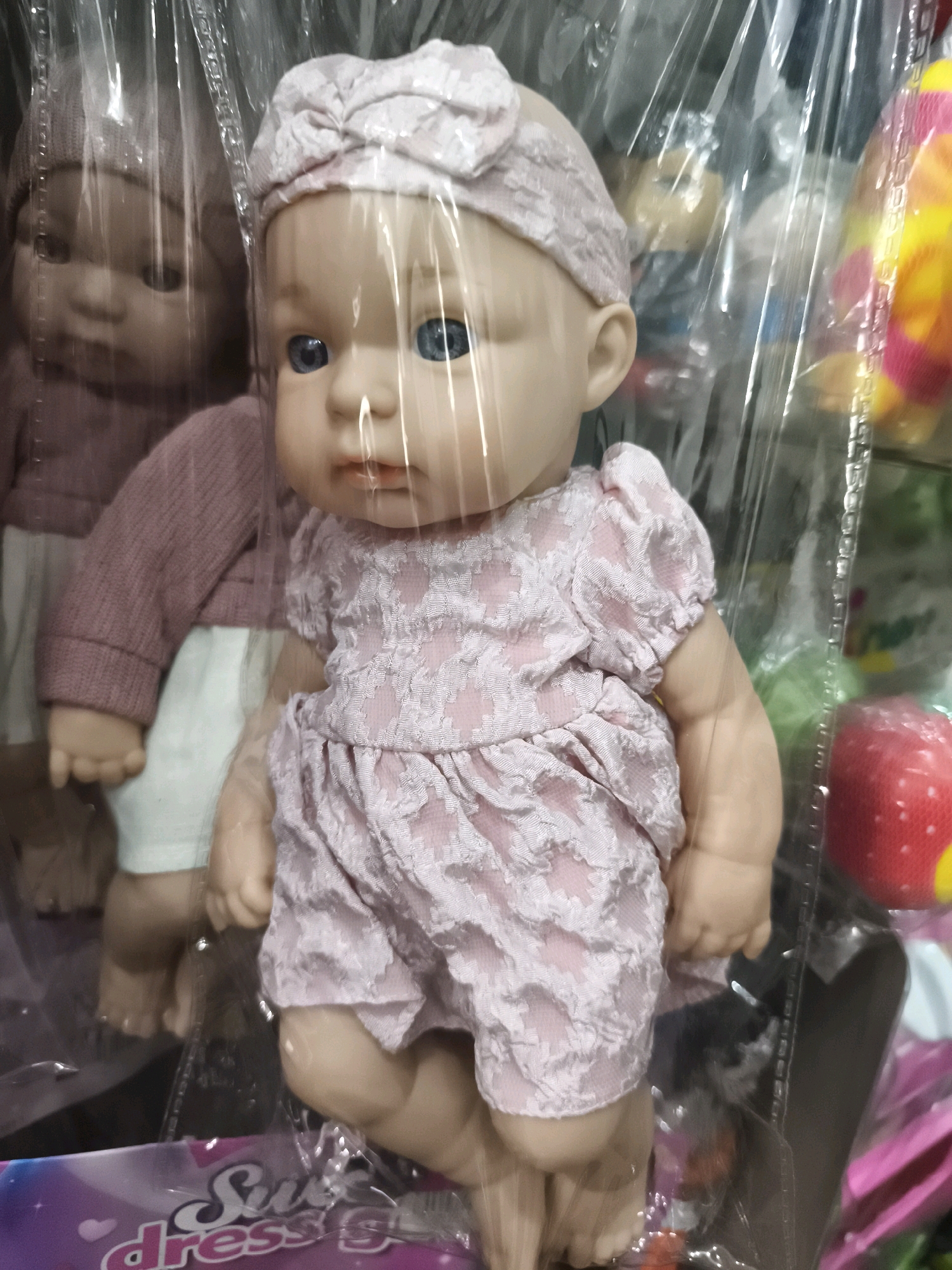 This is a close-up, eye-level, indoor photograph of two baby dolls in a retail setting, possibly a toy store or a market stall in Palembang, Indonesia. The dolls are encased in clear plastic packaging, which creates reflections and some blurring.
The foreground doll is a Caucasian-style baby doll with blue eyes and fair skin. It is wearing a pink, textured romper with puffed sleeves and a matching headband with a bow. The doll has its head tilted slightly to the left, and its gaze is directed forward. Its hands are positioned in its lap. The doll's legs are bent.
Behind the foreground doll, partially obscured by the packaging and the front doll, is another doll. This doll appears to have darker skin and is wearing a pink knitted sweater over a white garment. Its face is visible, with dark eyes.
The background is cluttered with other items, including what appear to be more toys or merchandise, some of which are brightly colored and appear to be wrapped in plastic or netting. The overall lighting suggests an indoor environment with artificial illumination. The image quality is somewhat compromised by the plastic packaging and reflections. The specific location in Palembang cannot be determined from the image alone, but the presence of the dolls and the retail environment suggest a commercial area.