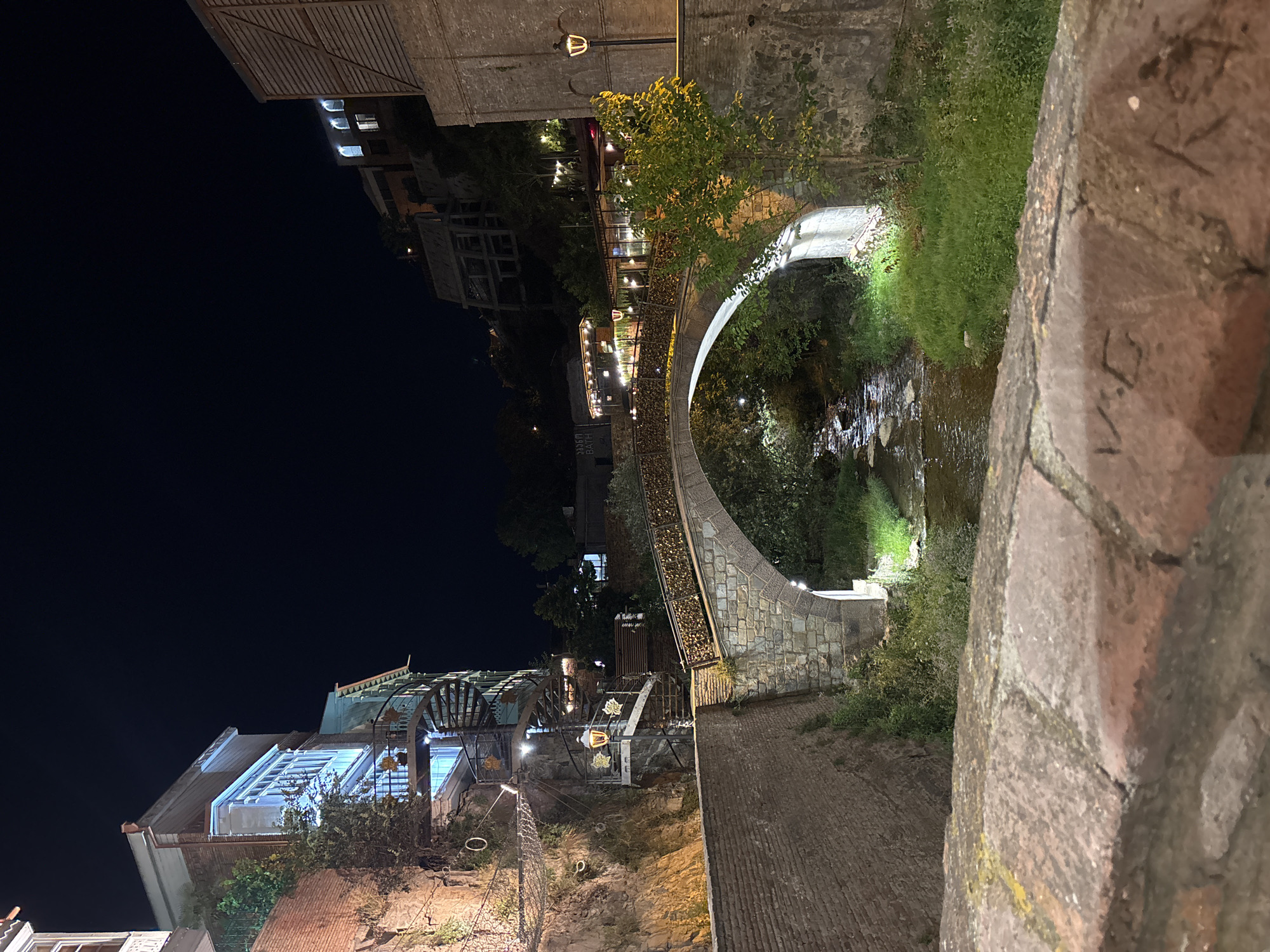 The image shows a nighttime view of a stone bridge spanning a narrow river. The bridge has a decorative railing adorned with numerous padlocks. On the left side of the image, a multi-story building with blue accents and spiral staircases is built into a rocky cliff. The building is illuminated by interior lights and external lamps. On the right side of the bridge, another building with wooden siding and illuminated windows is visible. The area around the riverbank is covered in grass and trees. The sky is dark, indicating it is nighttime. A brick wall is in the foreground, with some inscriptions visible on its surface.