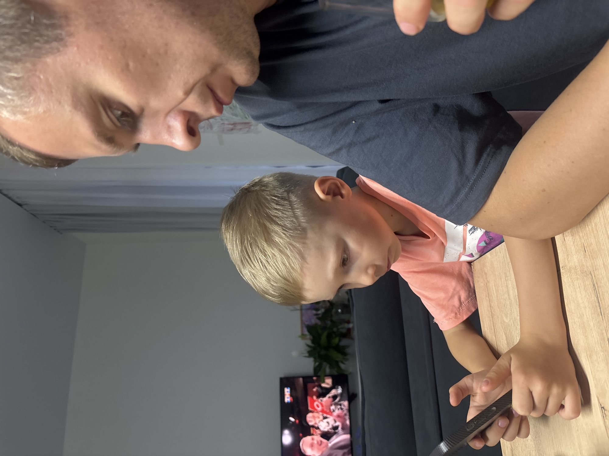 Within a residential setting in Chaikovsky, Russia, an adult male and a young male child are seated at a light-colored wooden table. The adult male, positioned on the right side of the frame, is seen in profile, looking downwards and to the left. He has short, light-colored hair, noticeable facial stubble, and wears a dark blue short-sleeved t-shirt with a prominent sweat stain under the right armpit. He holds a clear glass containing a yellowish liquid in his right hand. To his left, the child, estimated to be between four and seven years old, is also in profile, intently focused on a dark-colored smartphone held with both hands. The child has short, very light blonde hair styled with a shaved design on the left side of the head and is wearing a peach-colored short-sleeved t-shirt with a graphic design. In the background, a flat-screen television displays content from the Russian channel "ТНТ," showing multiple individuals, one prominently in a suit. A dark gray sofa, potted plants, and light-colored walls with sheer white curtains complete the domestic interior.